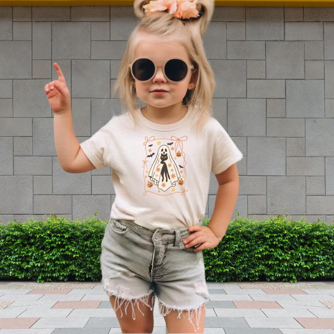 A young girl wearing a white t-shirt with a graphic design, sunglasses, and denim shorts is standing in front of a brick wall.