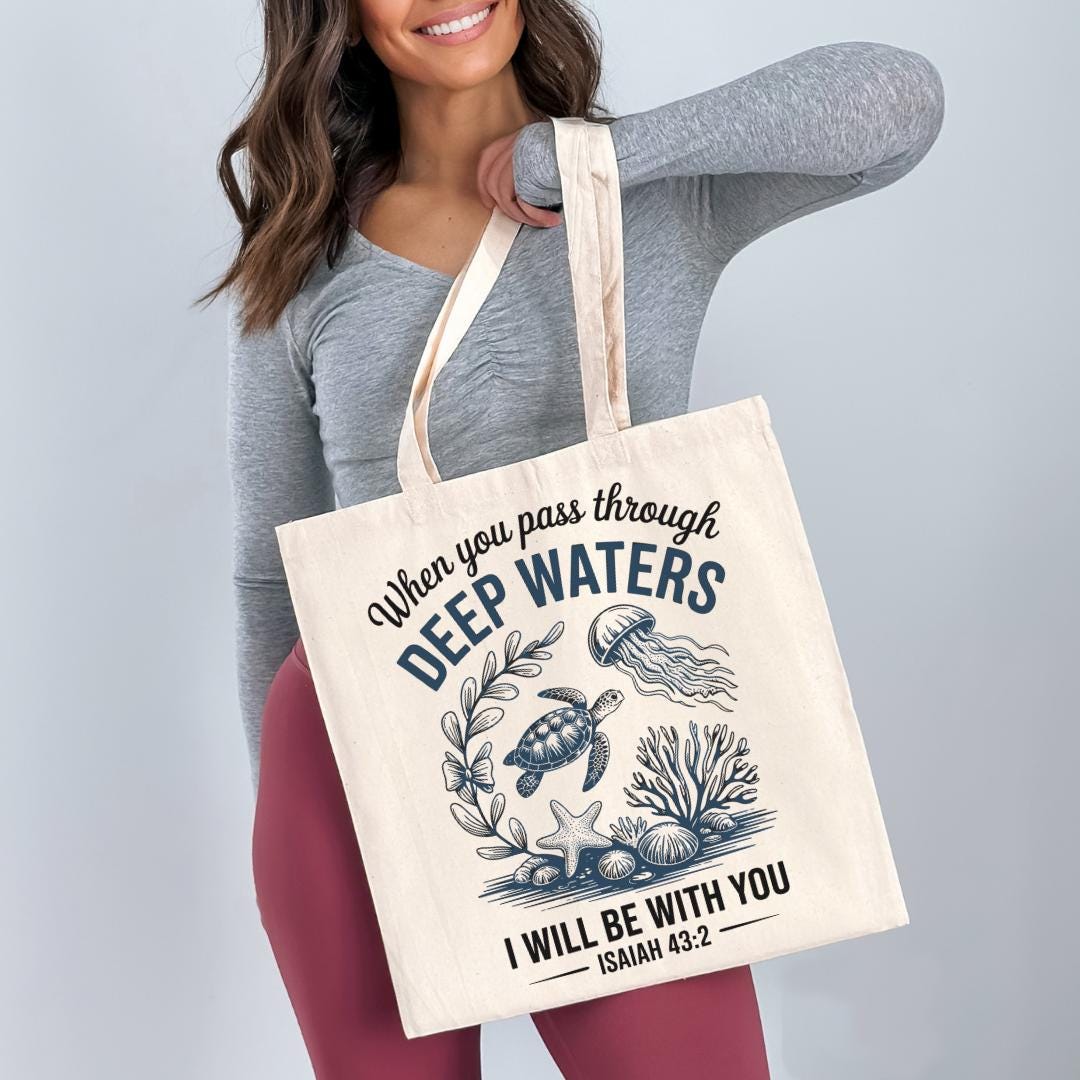 A woman is holding a white tote bag with a printed design of marine life and text that reads "When you pass through DEEP WATERS, I WILL BE WITH YOU" and " Isaiah 43:2".