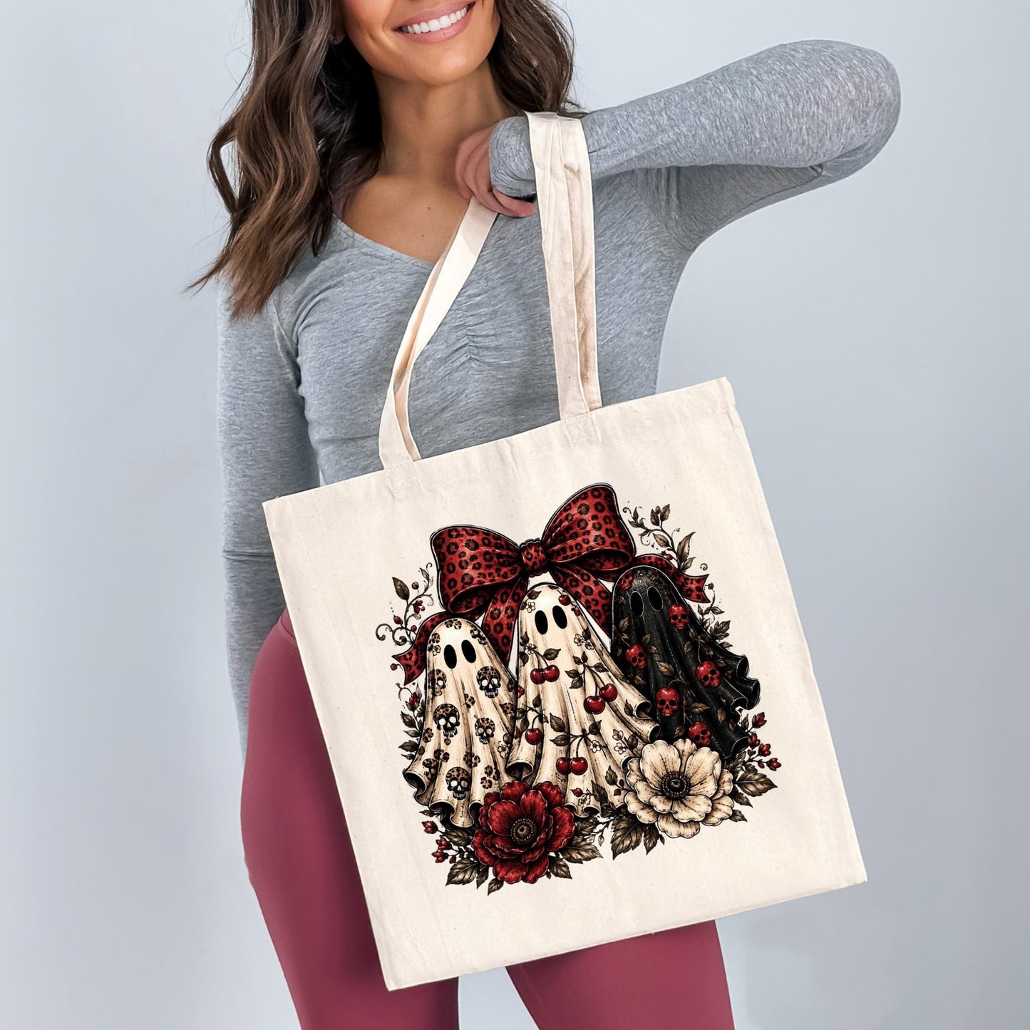 A woman is holding a white tote bag with a graphic design featuring three ghostly figures surrounded by flowers and a red bow.