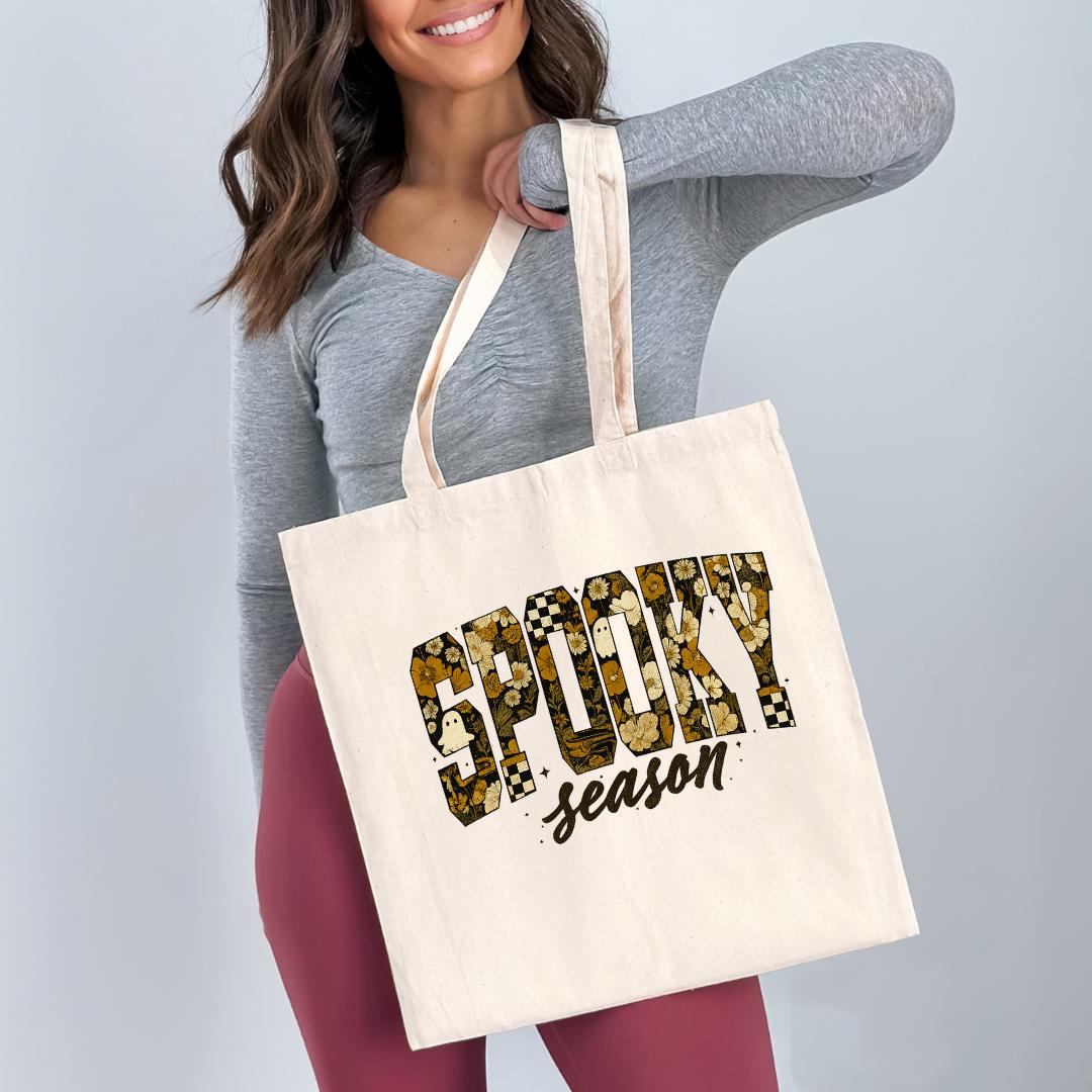 A woman is holding a white tote bag with the text "SPOOKY season" printed on it.
