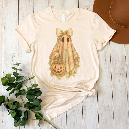 A white t-shirt with a Halloween-themed graphic of a ghost holding a pumpkin and a bow, accompanied by a brown hat and some greenery on a wooden surface.
