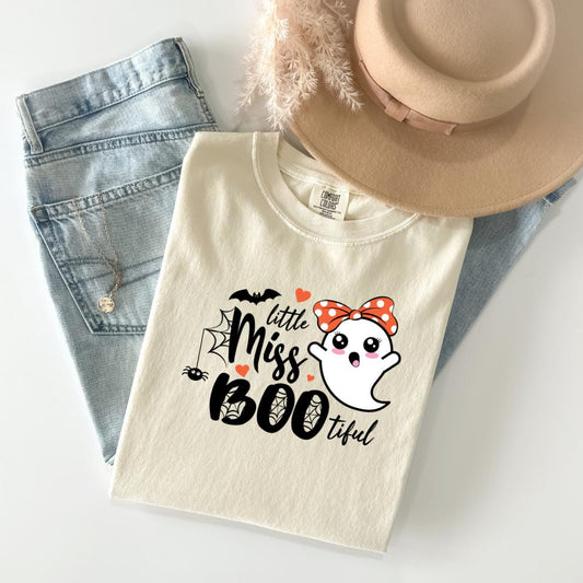 A white t-shirt with a graphic design featuring a ghost character and the text "Little Miss Boo" printed on it, along with a bow and a spider web. The t-shirt is placed on top of a pair of jeans and a beige hat.