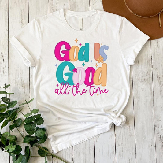 A white t-shirt with a colorful graphic design that says "God is good all the time" printed on it.