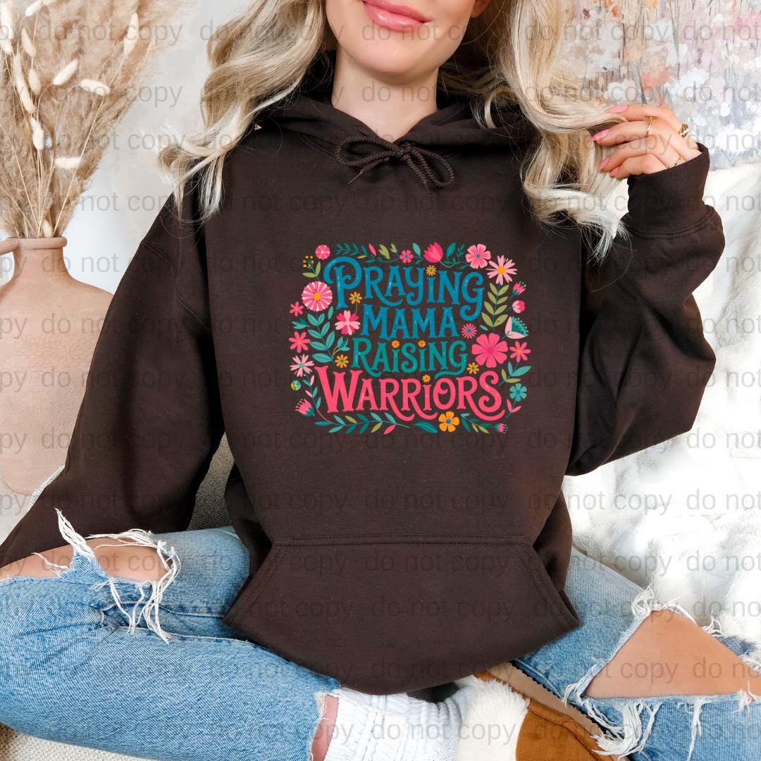 A woman wearing a brown hoodie with a colorful graphic design that says "Praying Mama Raising Warriors" printed on it.