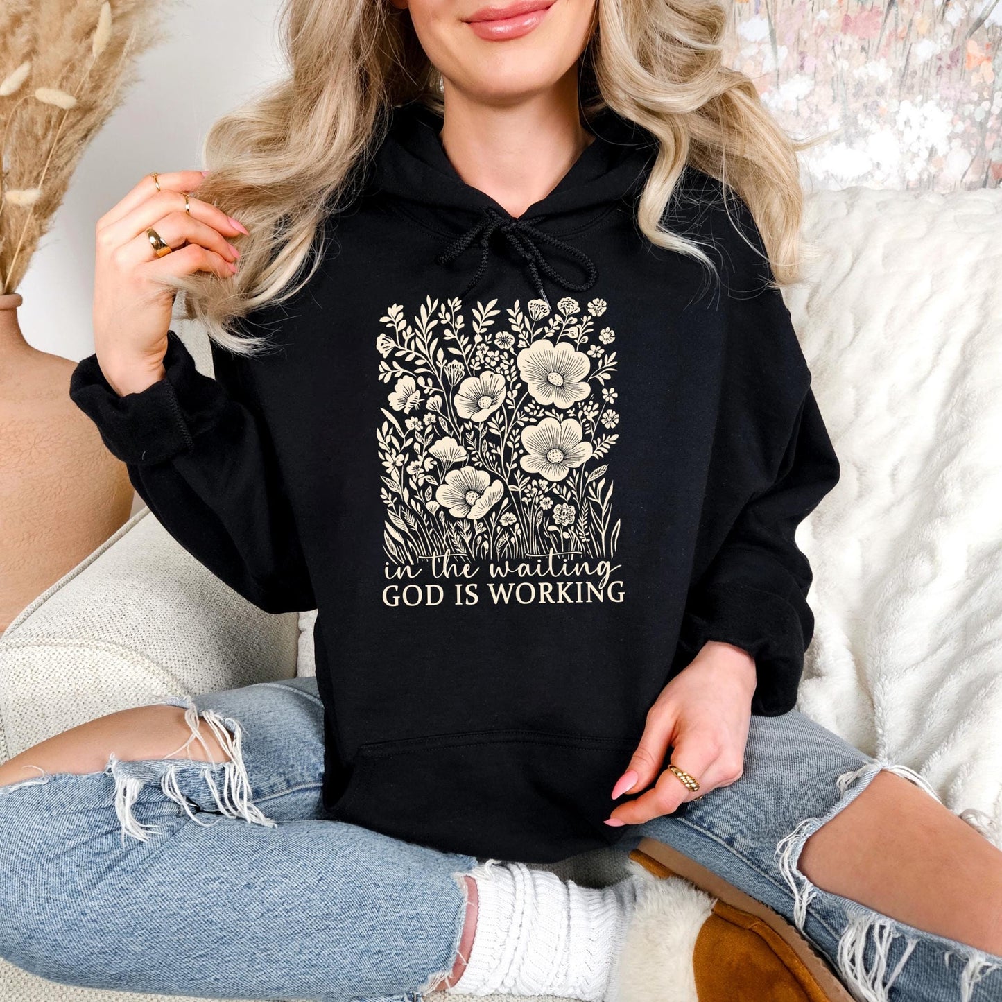 A woman wearing a black hoodie with a floral design and the text "in the waiting God is working" is sitting on a couch.