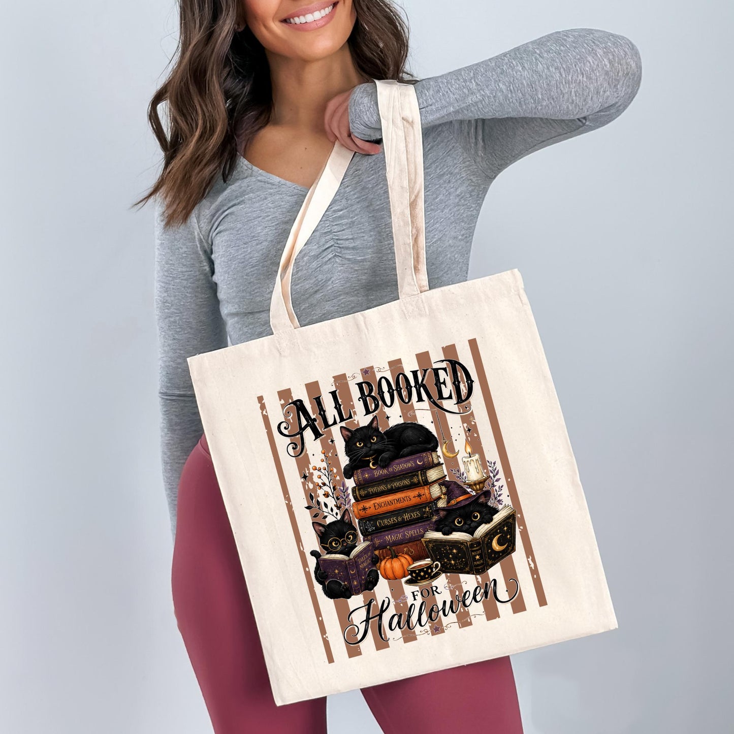 A woman is holding a white tote bag with a printed design of cats and books, along with the text "All Booked for Halloween".