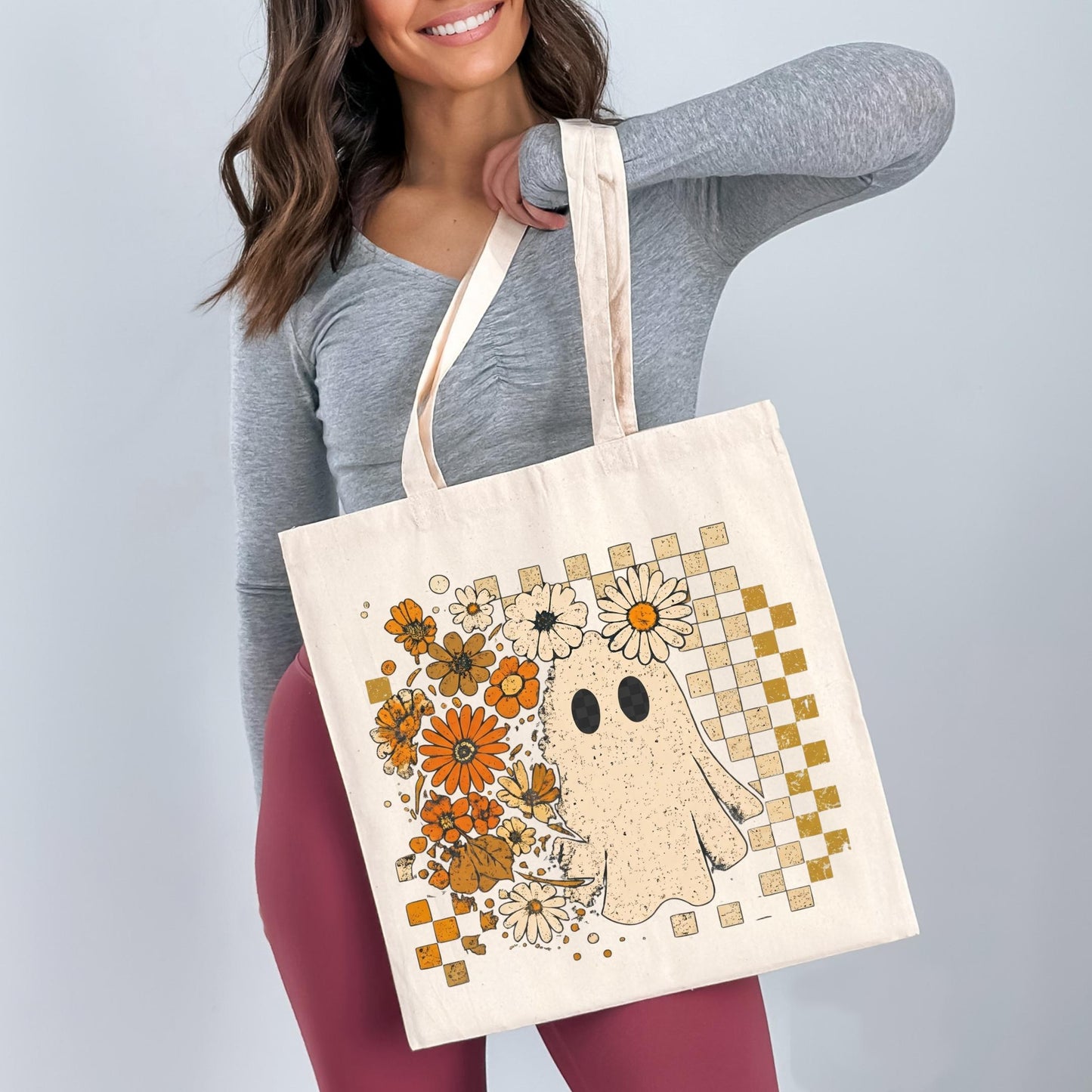A woman is holding a large canvas tote bag with a ghost design on it, surrounded by flowers.