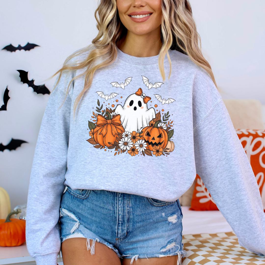 A woman wearing a grey sweatshirt with a graphic of a ghost, pumpkins, and flowers, standing in front of a Halloween-themed background.