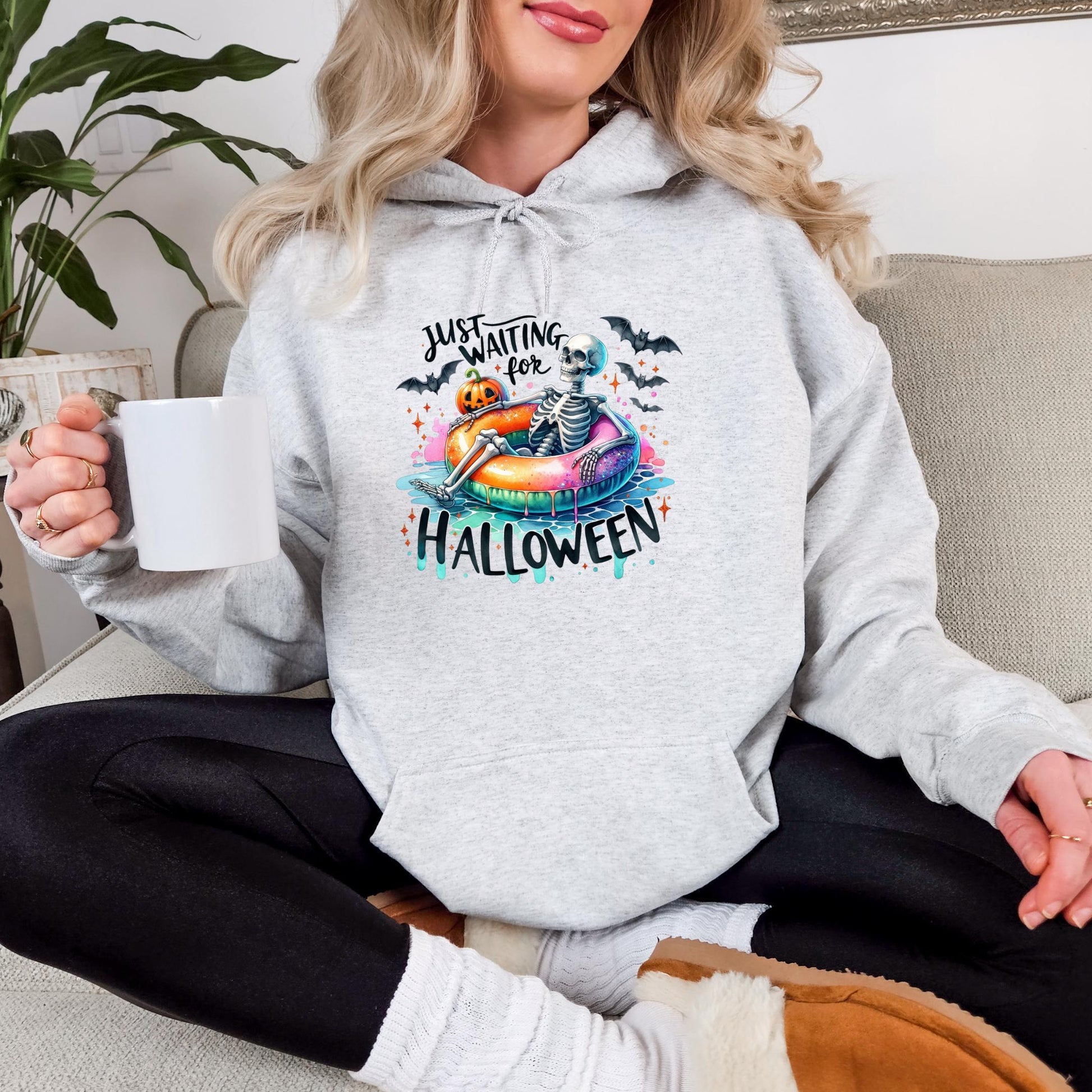 A woman wearing a grey hoodie with a Halloween-themed graphic and text, sitting on a couch and holding a white mug.