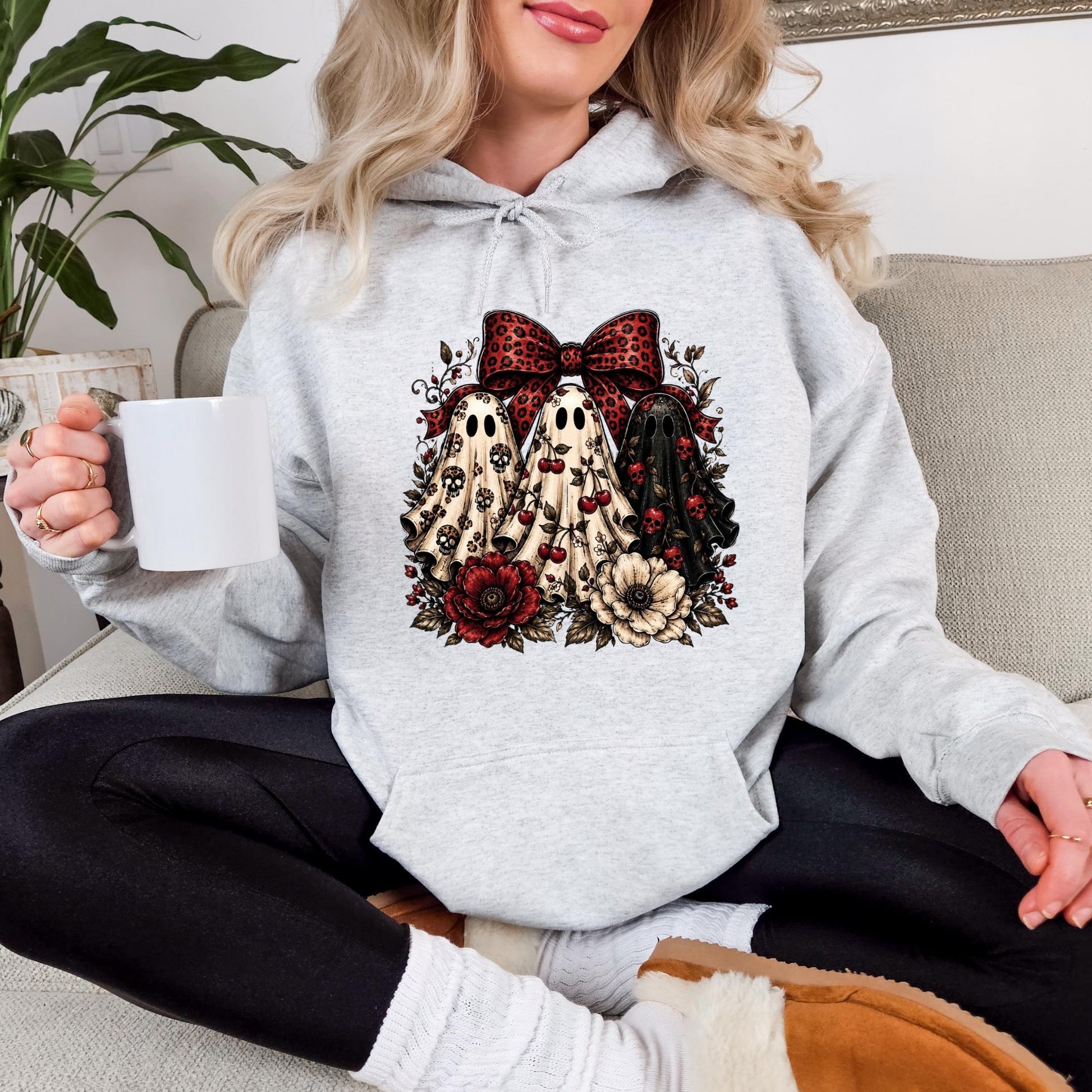 A woman wearing a grey hoodie with a graphic design of three ghostly figures and floral elements. She is sitting on a couch and holding a white mug.