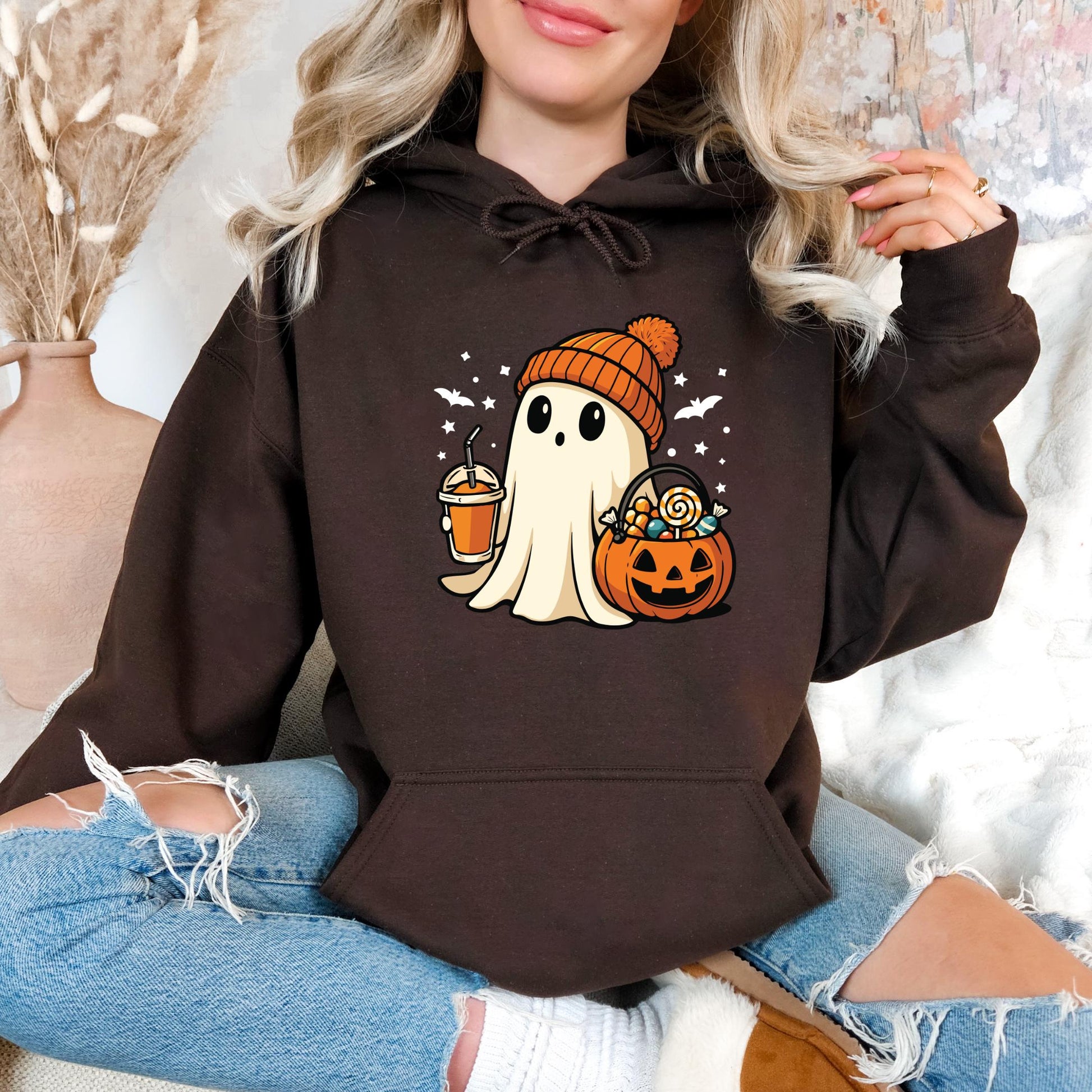 A woman wearing a brown hoodie with a Halloween-themed graphic of a ghost holding a pumpkin and a drink.
