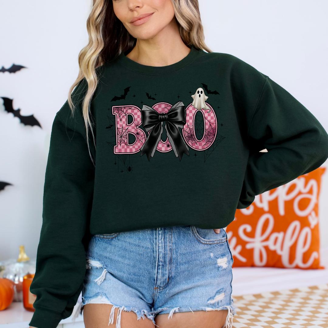 A woman wearing a green sweatshirt with the word "BOO" printed on it, along with a bow and a ghost. She is also wearing denim shorts and a black top.