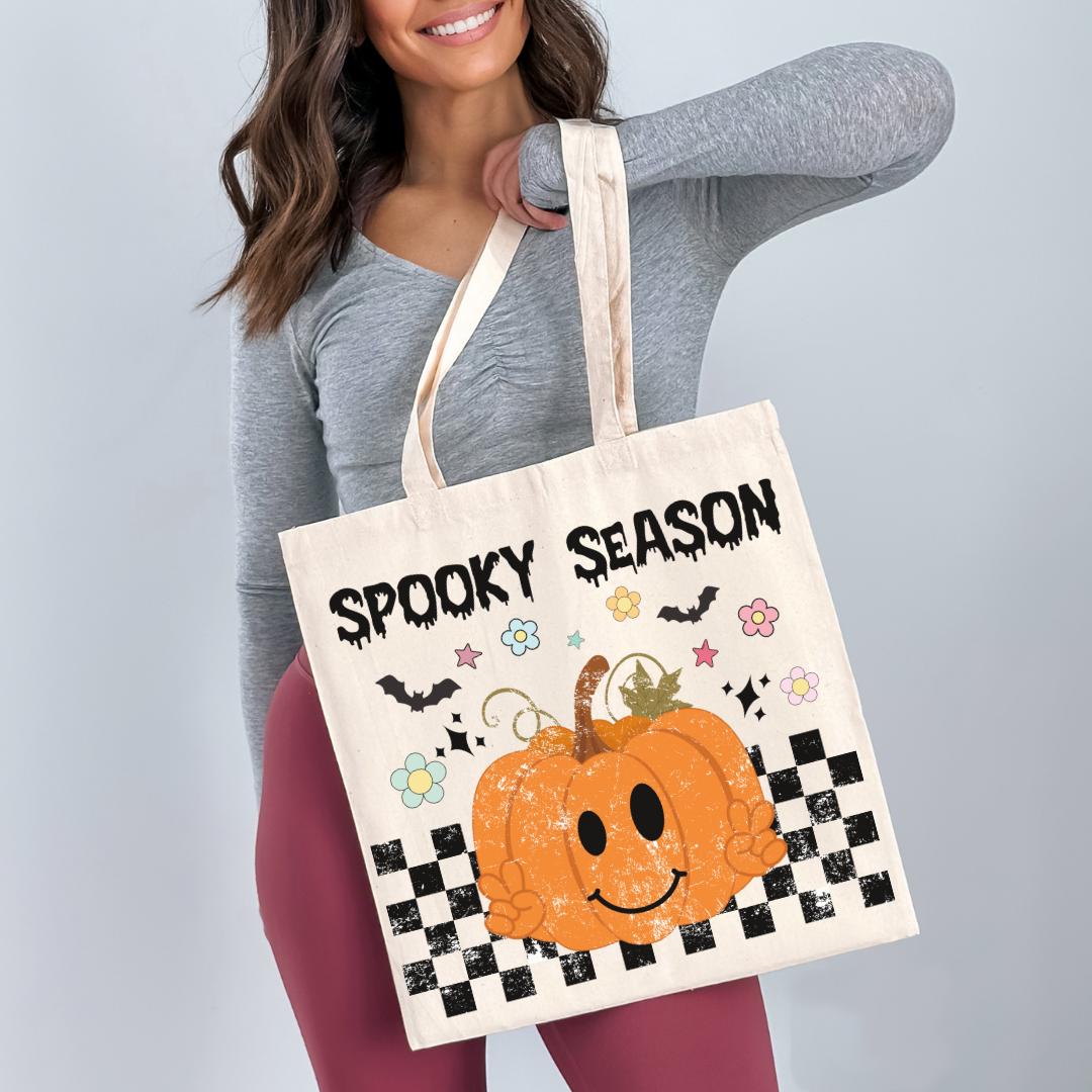 A woman is holding a white tote bag with a pumpkin and Halloween-themed design.