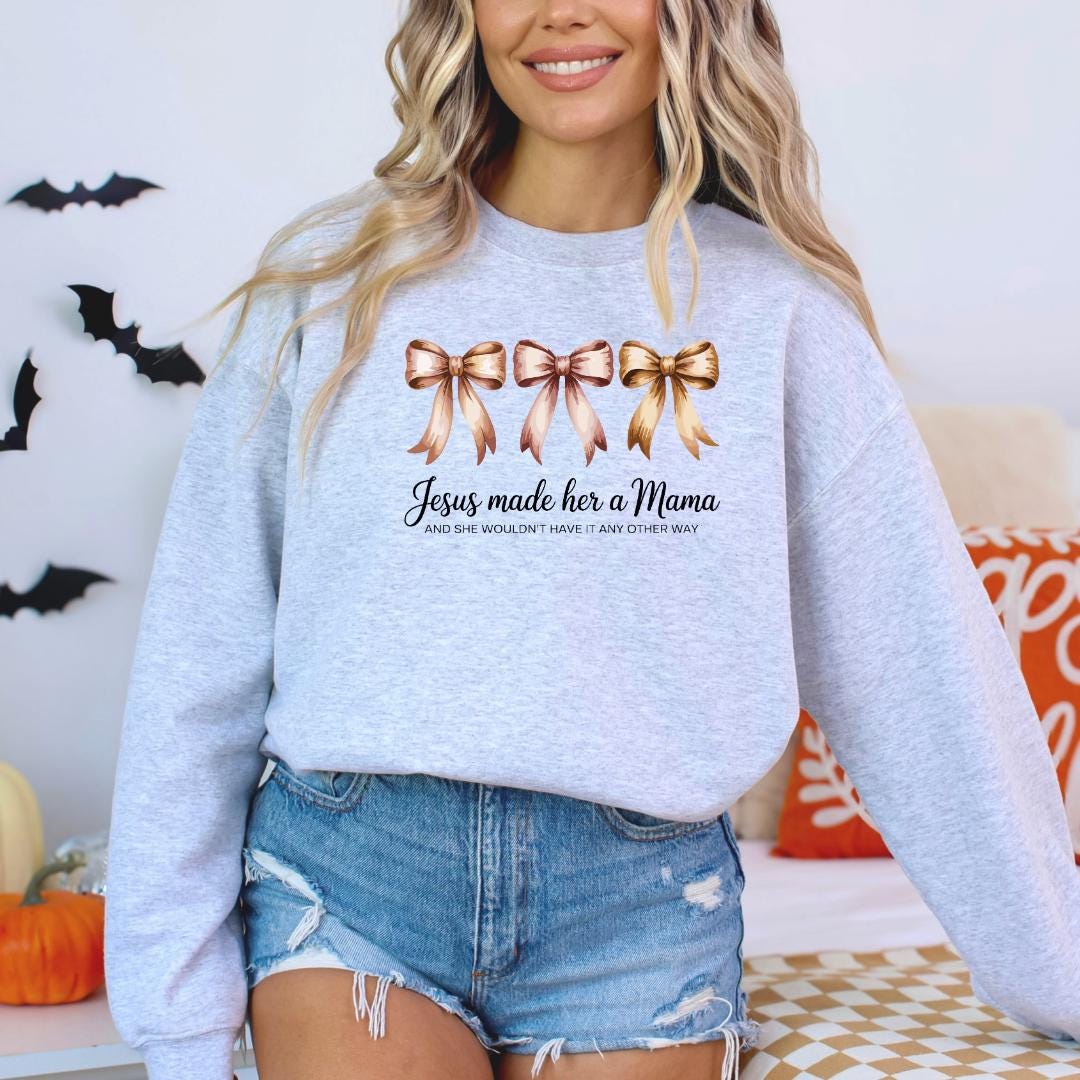 A woman wearing a gray sweatshirt with a graphic design of three bows and text that reads "Jesus made her a Mama and she wouldn't have it any other way".