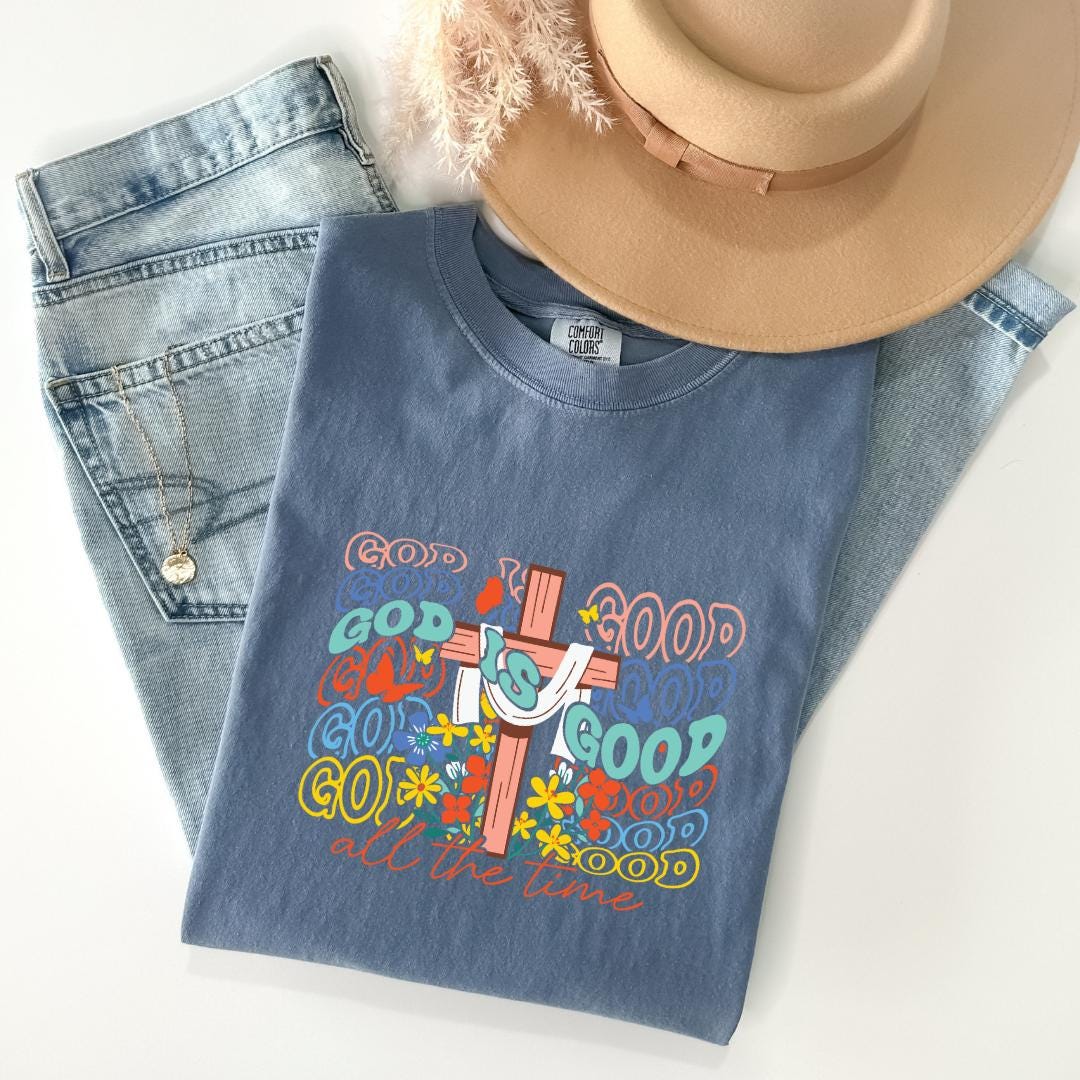A blue t-shirt with a colorful graphic design, a pair of jeans, and a beige hat are arranged on a white surface.