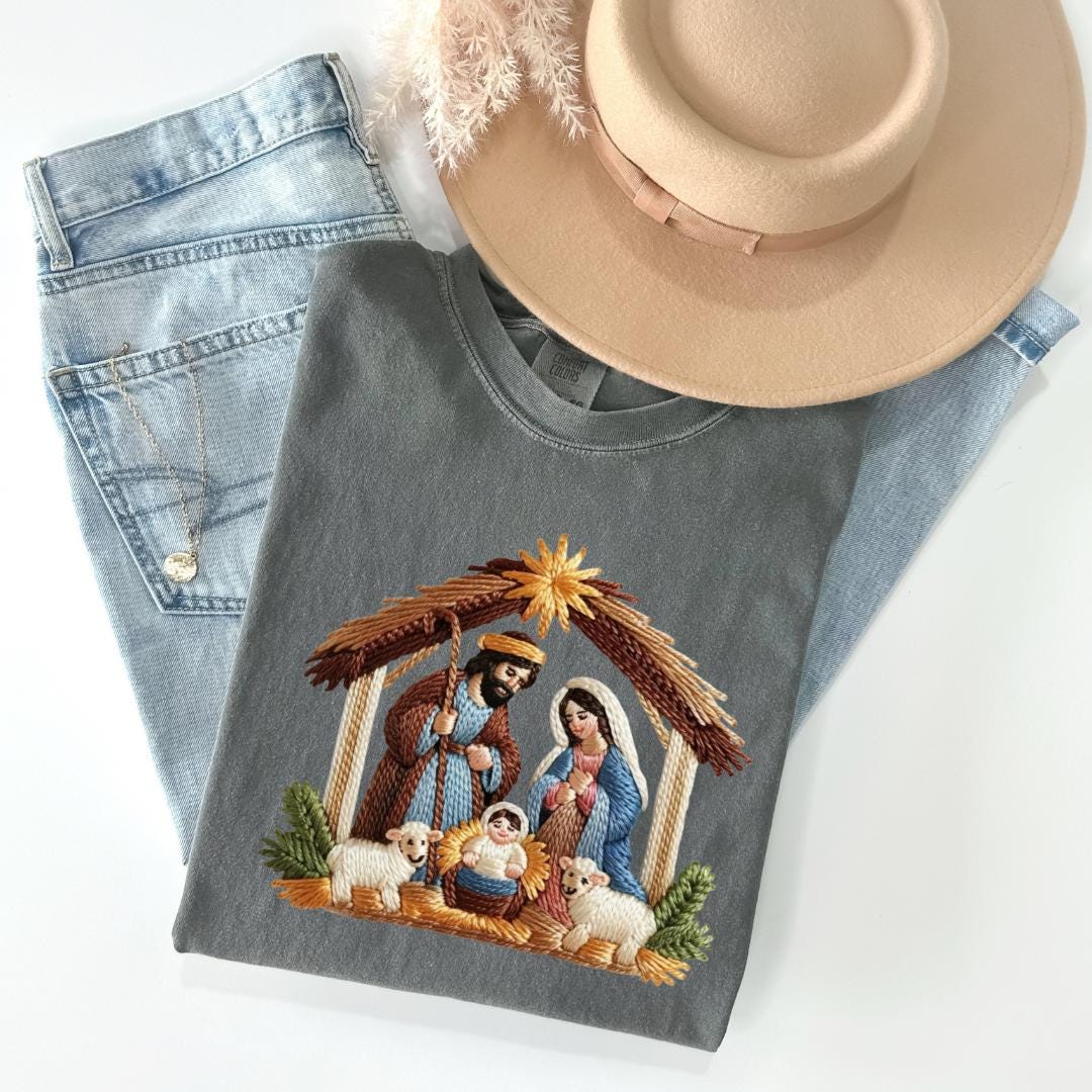 A gray t-shirt with an embroidered nativity scene, a pair of blue jeans, and a beige hat are arranged on a white background.