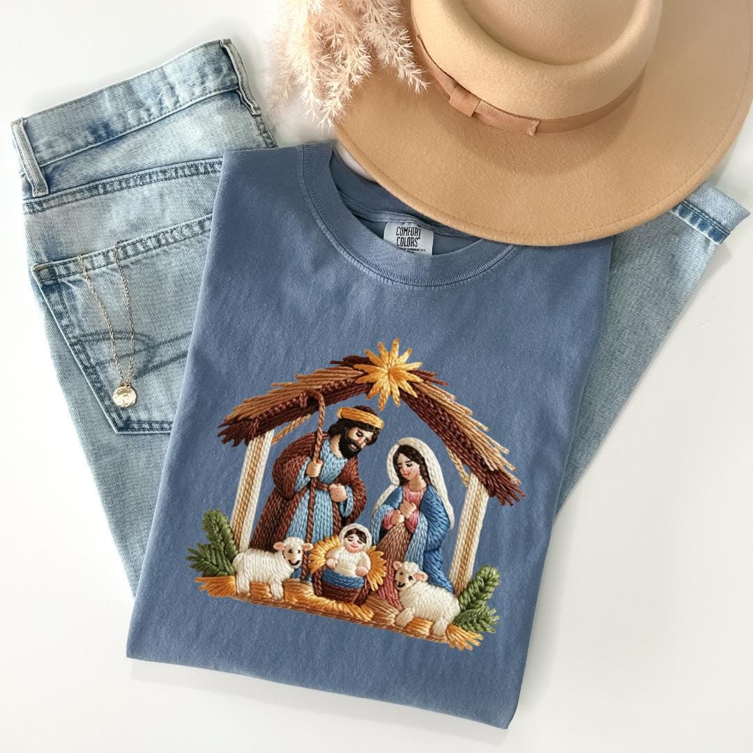 A blue t-shirt with an embroidered image of a nativity scene, featuring a man, a woman, a baby, and animals, is displayed alongside a pair of jeans and a beige hat.