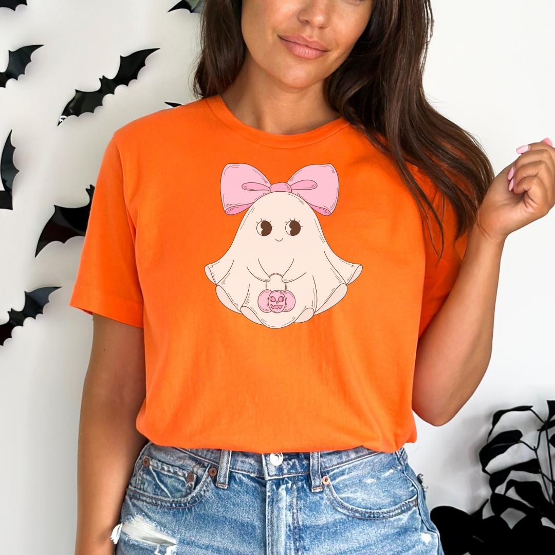 A woman wearing an orange t-shirt with a cartoon ghost and a pink bow on it, standing in front of a wall with black bats.