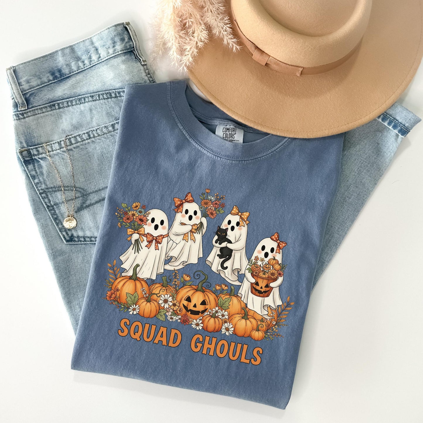 A blue t-shirt with a graphic of four ghostly figures holding pumpkins and flowers, along with the text "SQUADGHOULS", is displayed on a white surface.