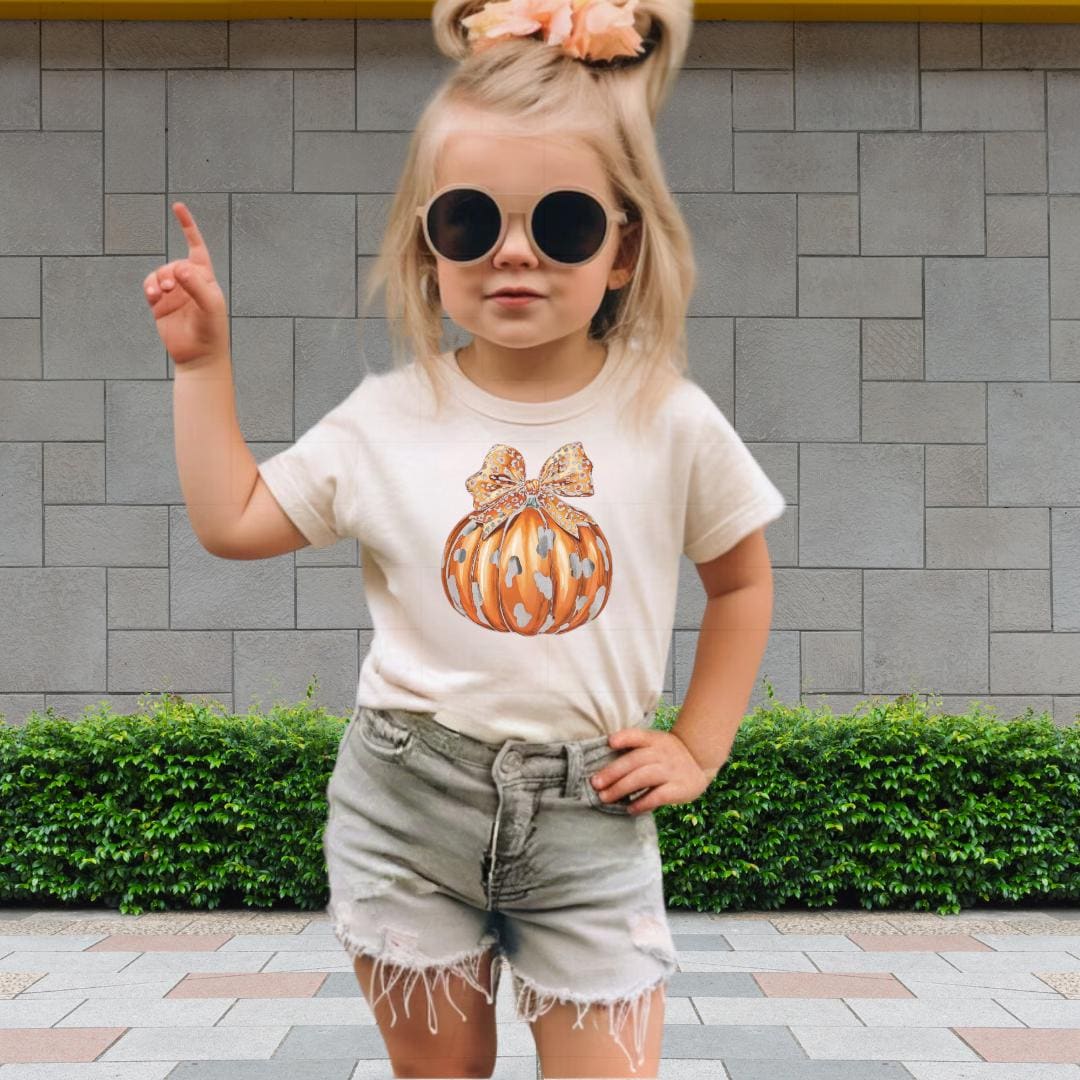 A young girl wearing a white t-shirt with a pumpkin graphic, sunglasses, and denim shorts is standing in front of a brick wall.