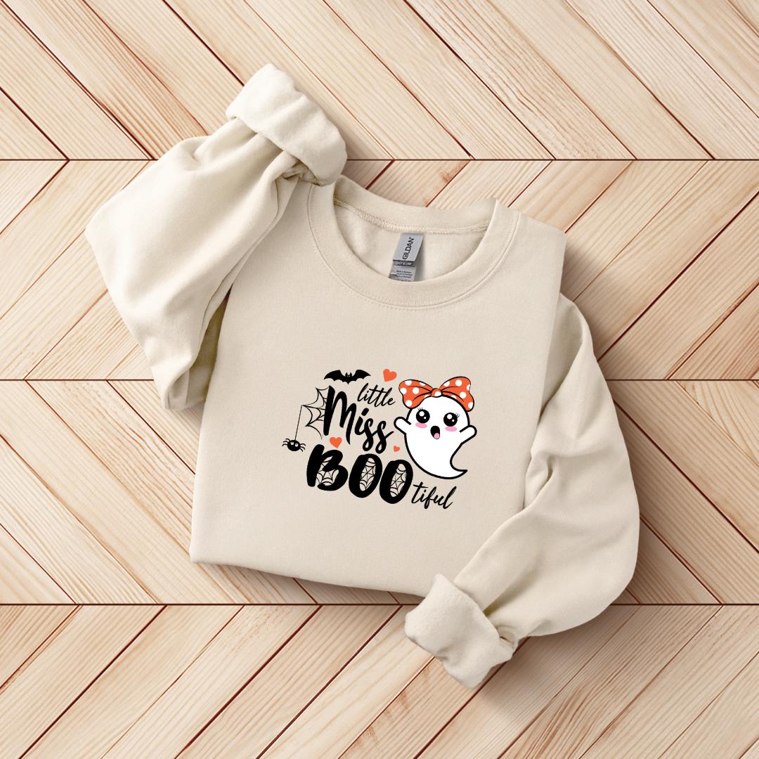 A beige sweater with a Halloween-themed graphic design, including a ghost character and the text "Little Miss Boook".