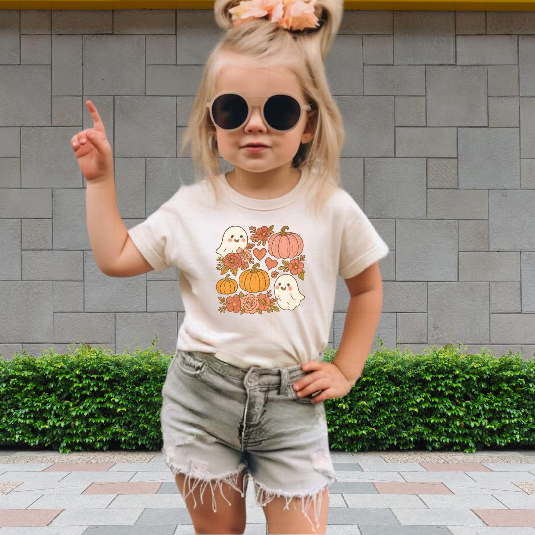 A young girl wearing a white t-shirt with a graphic of ghostly figures and pumpkins, sunglasses, and denim shorts is standing in front of a brick wall.