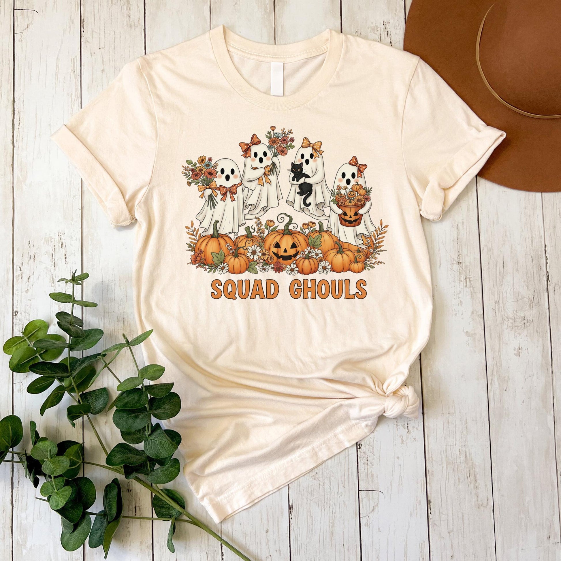 A white t-shirt with a graphic design featuring a group of ghostly figures surrounded by pumpkins and flowers, along with the text "SQUADGHOLS" printed on it.