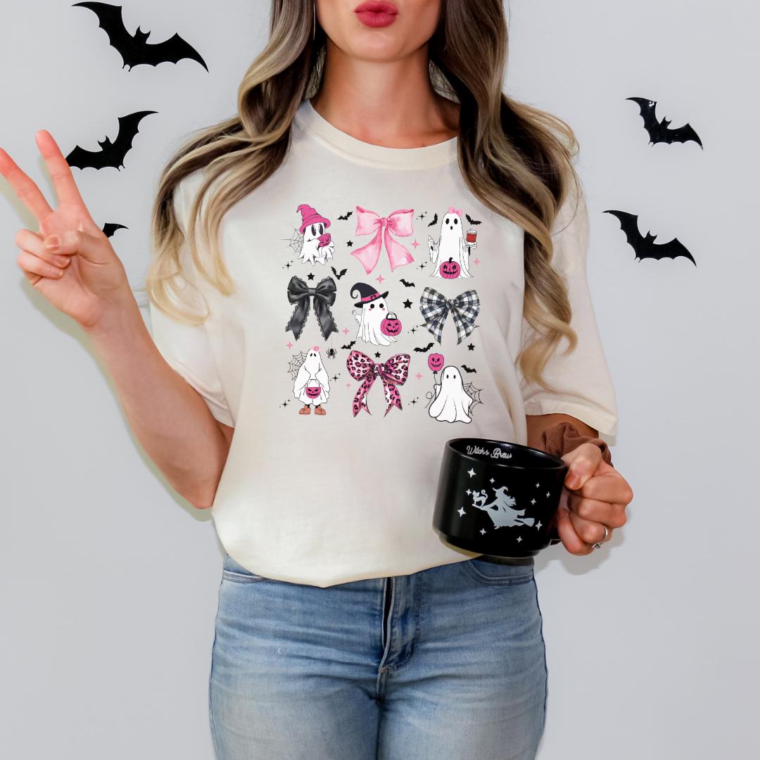 A woman is standing against a white background, wearing a white t-shirt with a graphic design featuring various Halloween-themed elements such as bats, ghosts, and bows. She is also holding a black mug with a similar design.