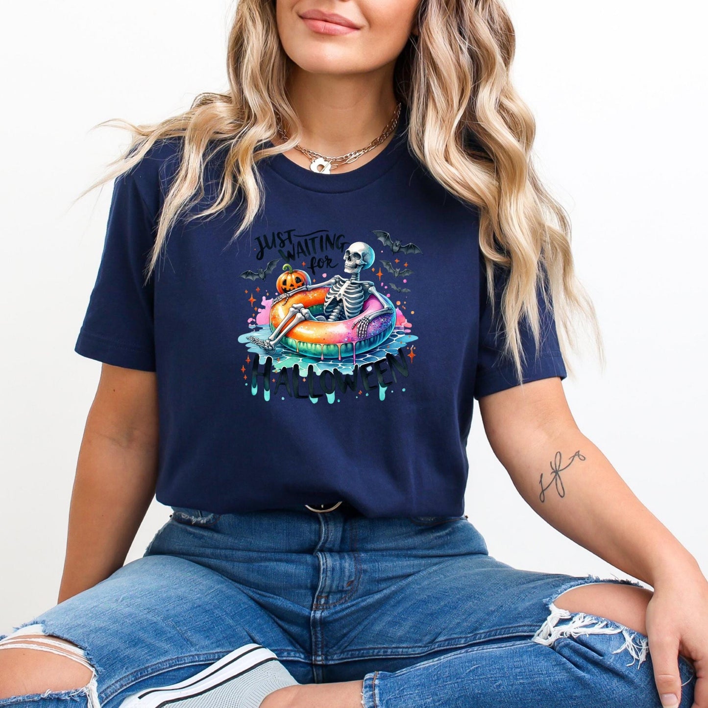A woman wearing a navy blue t-shirt with a graphic design of a skeleton on a donut, sitting on a beach.
