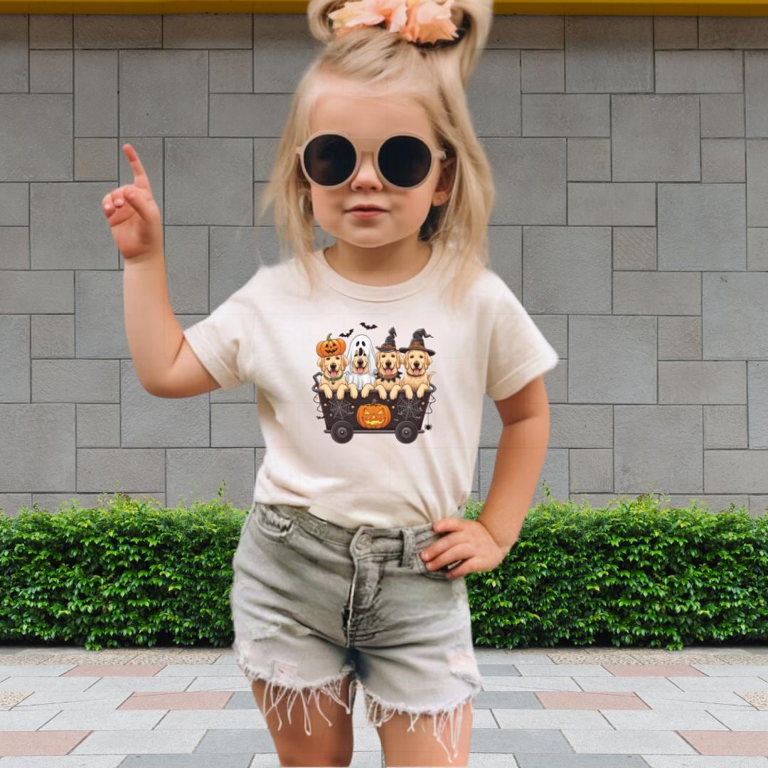 A young girl wearing a white t-shirt with a Halloween-themed graphic, sunglasses, and denim shorts is standing in front of a brick wall.