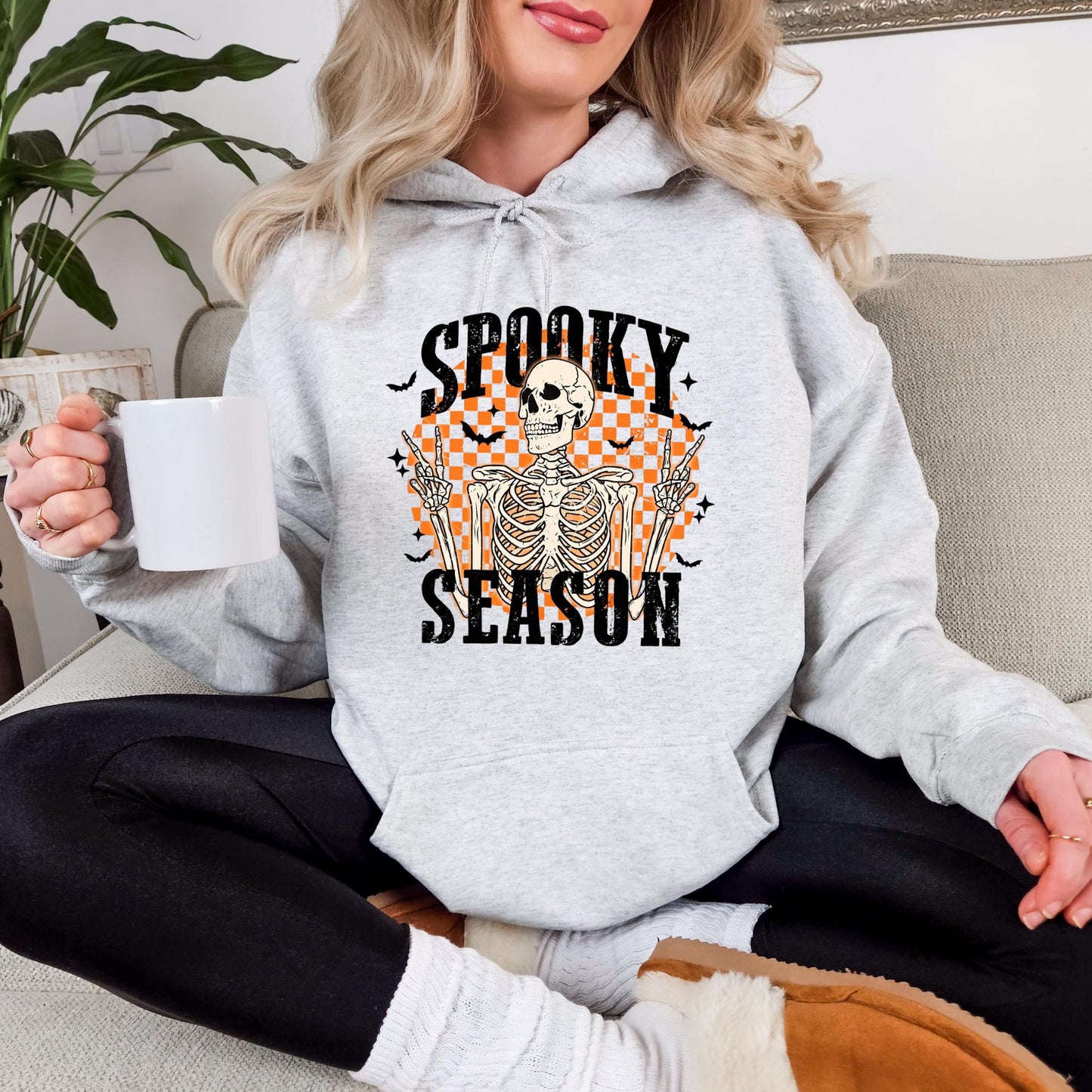 A woman wearing a grey hoodie with a Halloween-themed graphic and text, sitting on a couch and holding a white mug.