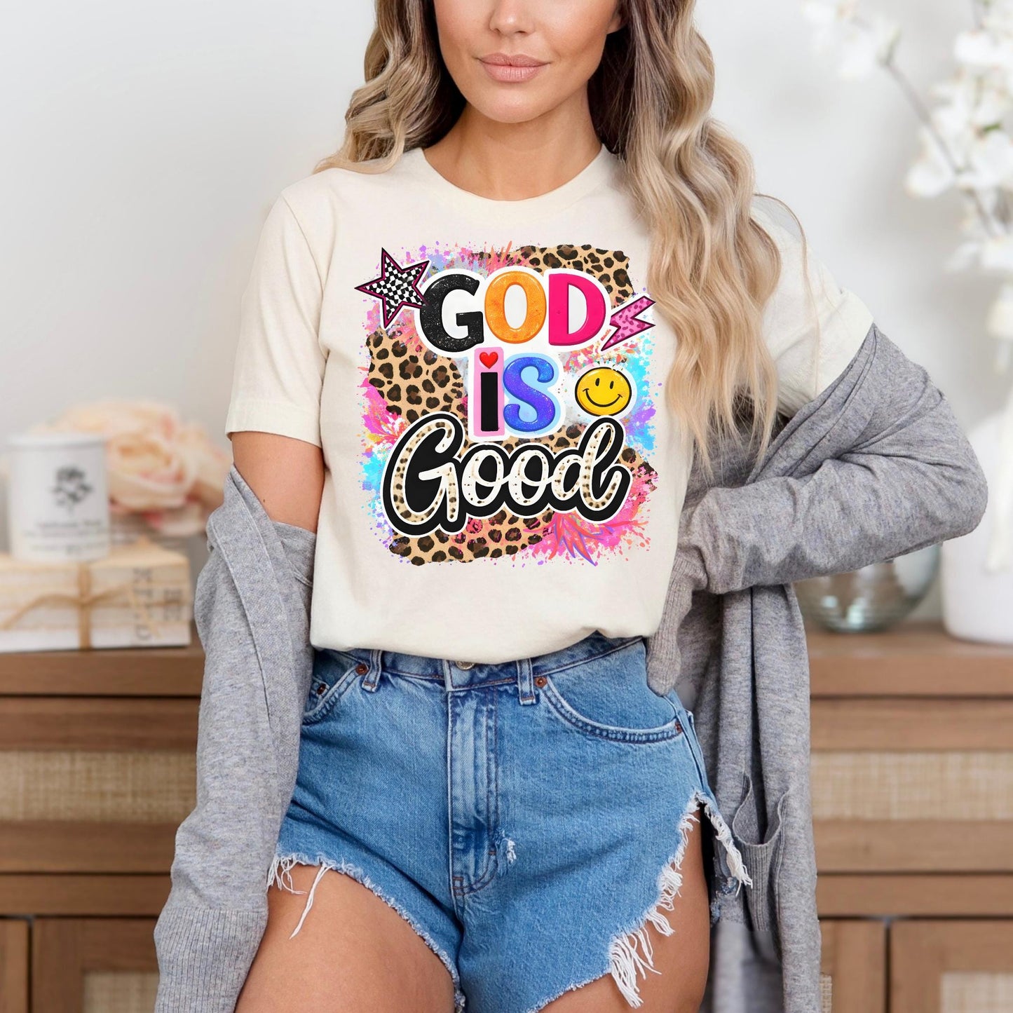 A woman wearing a white t-shirt with a colorful graphic design and denim shorts stands in front of a wooden cabinet.