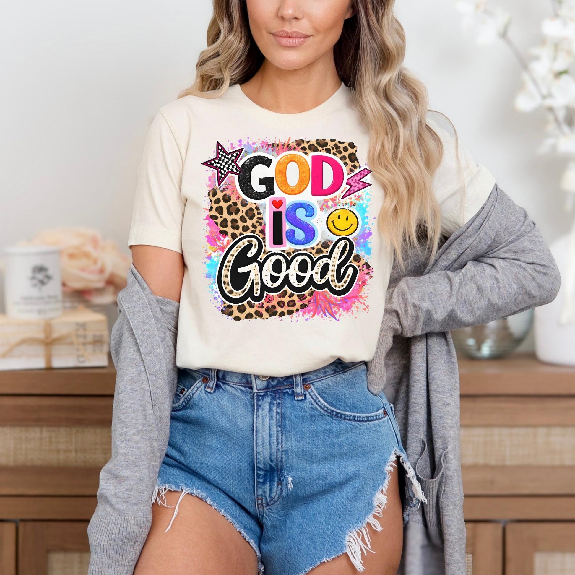 A woman wearing a white t-shirt with a colorful graphic design and denim shorts stands in front of a wooden cabinet.