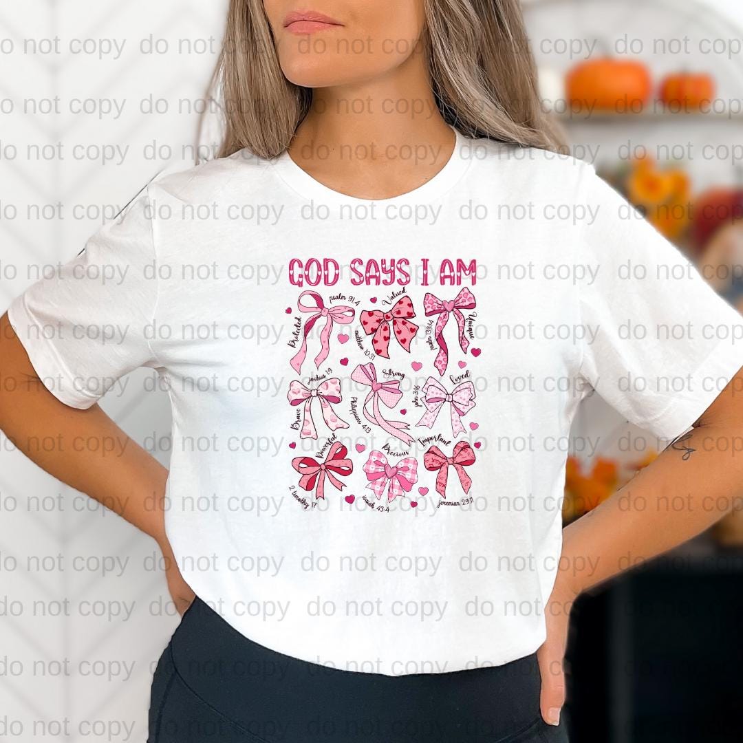A woman wearing a white t-shirt with a graphic design featuring pink bows and the text "God says I am" printed on it.