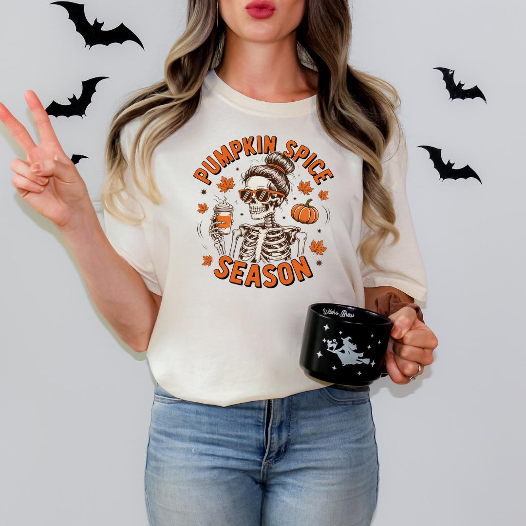 A woman wearing a white t-shirt with a pumpkin spice season graphic and holding a black mug with a skull design.