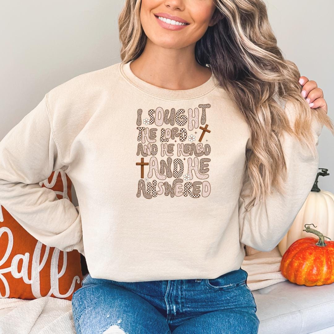 A woman wearing a beige sweatshirt with a text design on it, smiling and posing in front of a pumpkin.