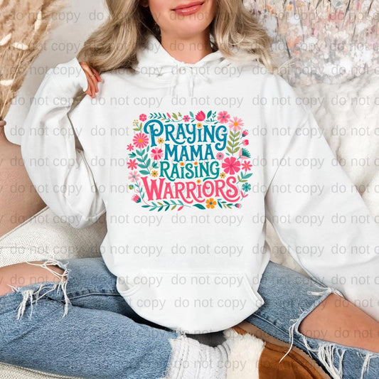 A woman wearing a white hoodie with a colorful graphic design that says "Praying Mama Raising Warriors".