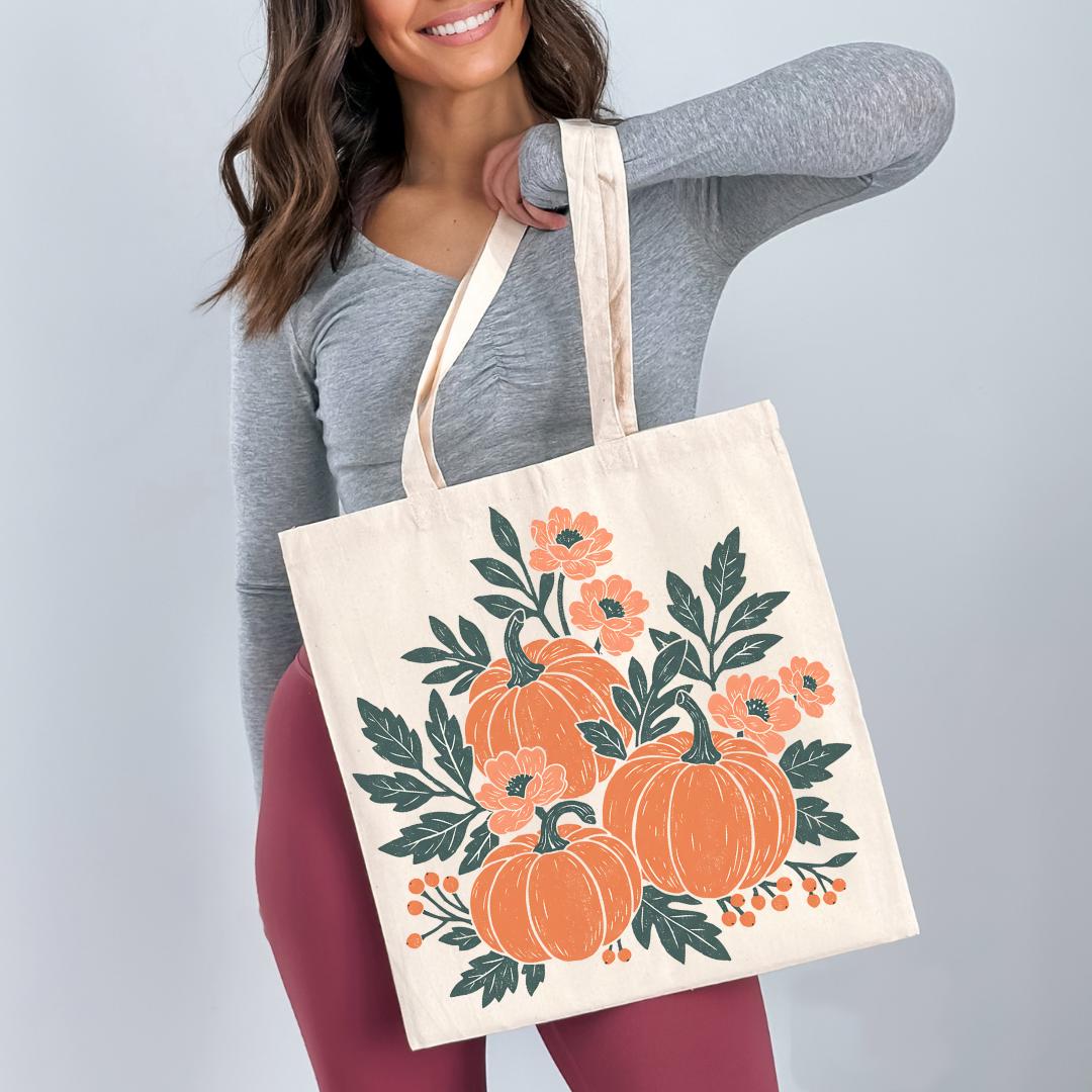 A woman is holding a white tote bag with a floral design featuring orange pumpkins and green leaves.