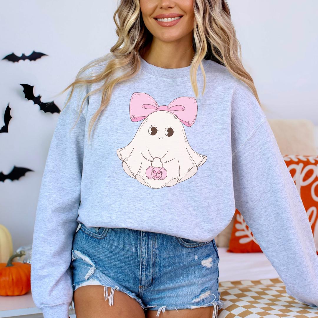A woman wearing a gray sweatshirt with a cartoon ghost design and a pink bow on her head, standing in front of a Halloween-themed background.