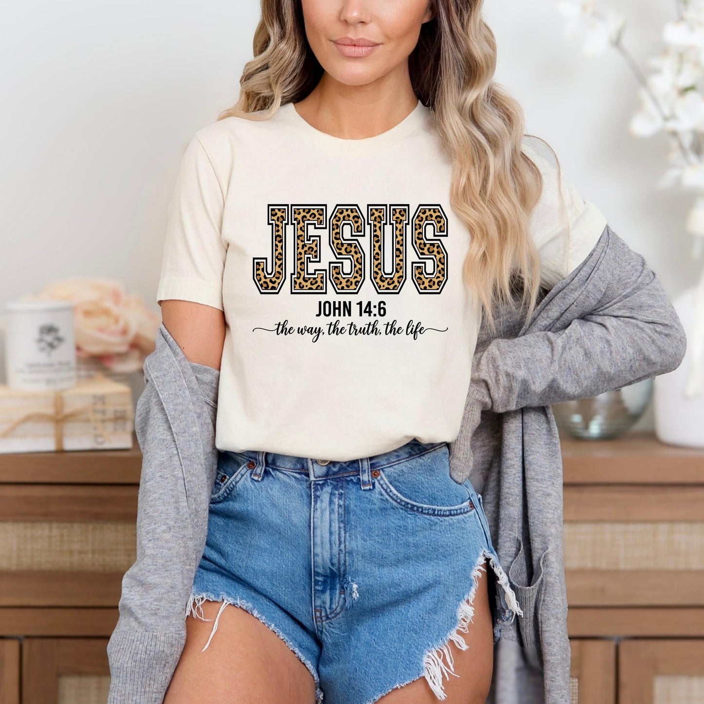 A woman wearing a white t-shirt with the text "JESUS" and "JOHN 14:6" printed on it, along with the phrase "the way, the truth, the life" below. She is also wearing denim shorts and a gray cardigan.