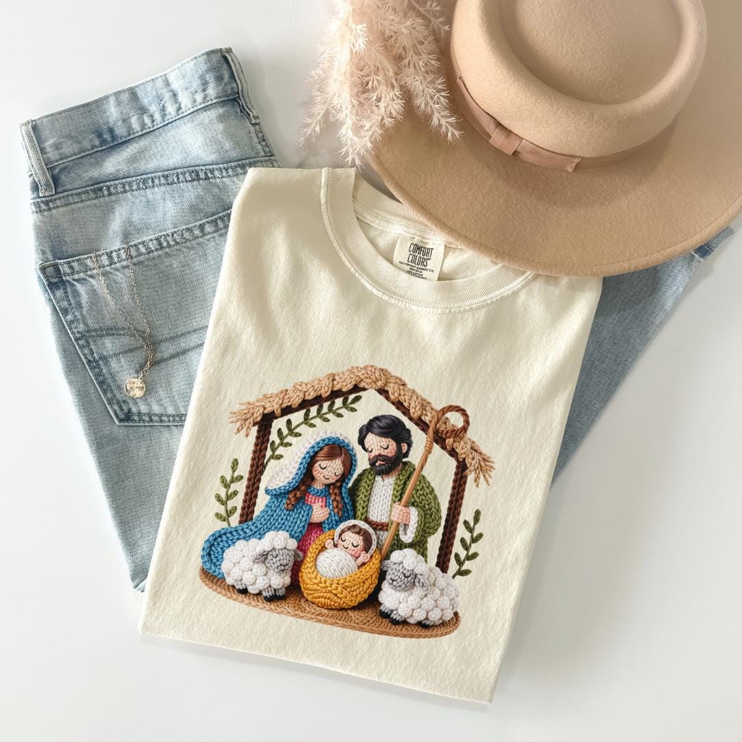 A white t-shirt with a crochet nativity scene design, a pair of blue jeans, and a beige hat are arranged on a white surface.