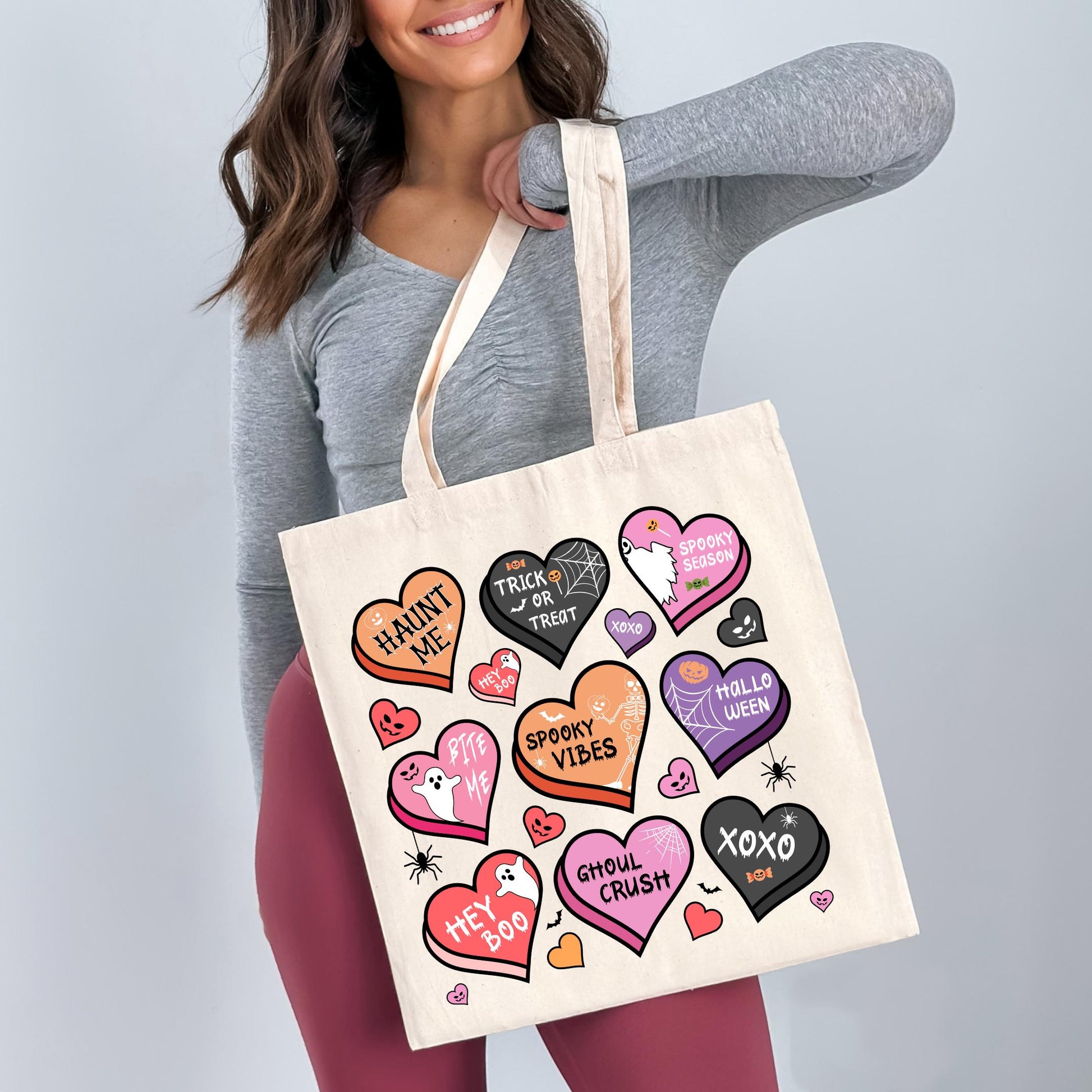 A woman is holding a white tote bag with various Halloween-themed heart designs and text printed on it.