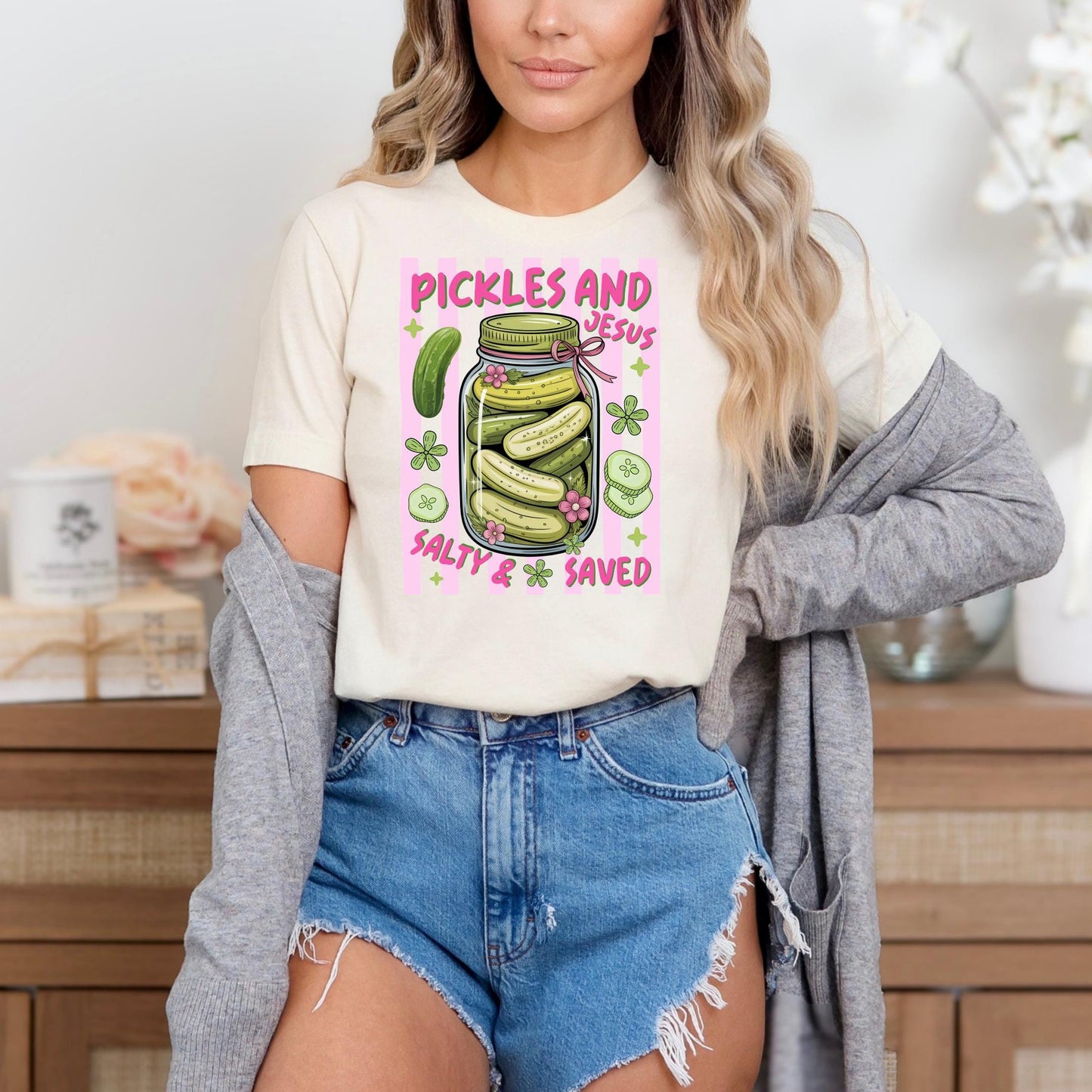 A woman wearing a white t-shirt with a graphic design of a jar of pickles and the text "Pickles and Jesus Salty & Saved" is standing in front of a wooden cabinet.
