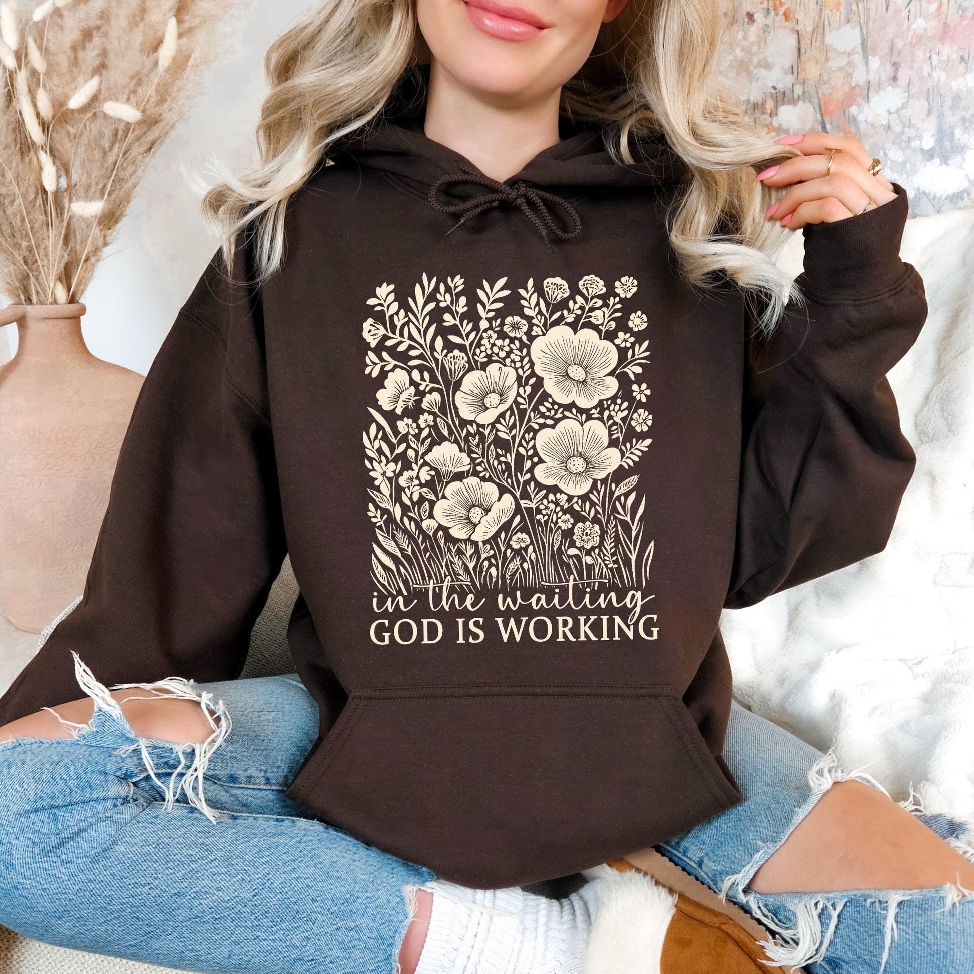 A woman wearing a brown hoodie with a floral design and text printed on it, sitting on a couch.