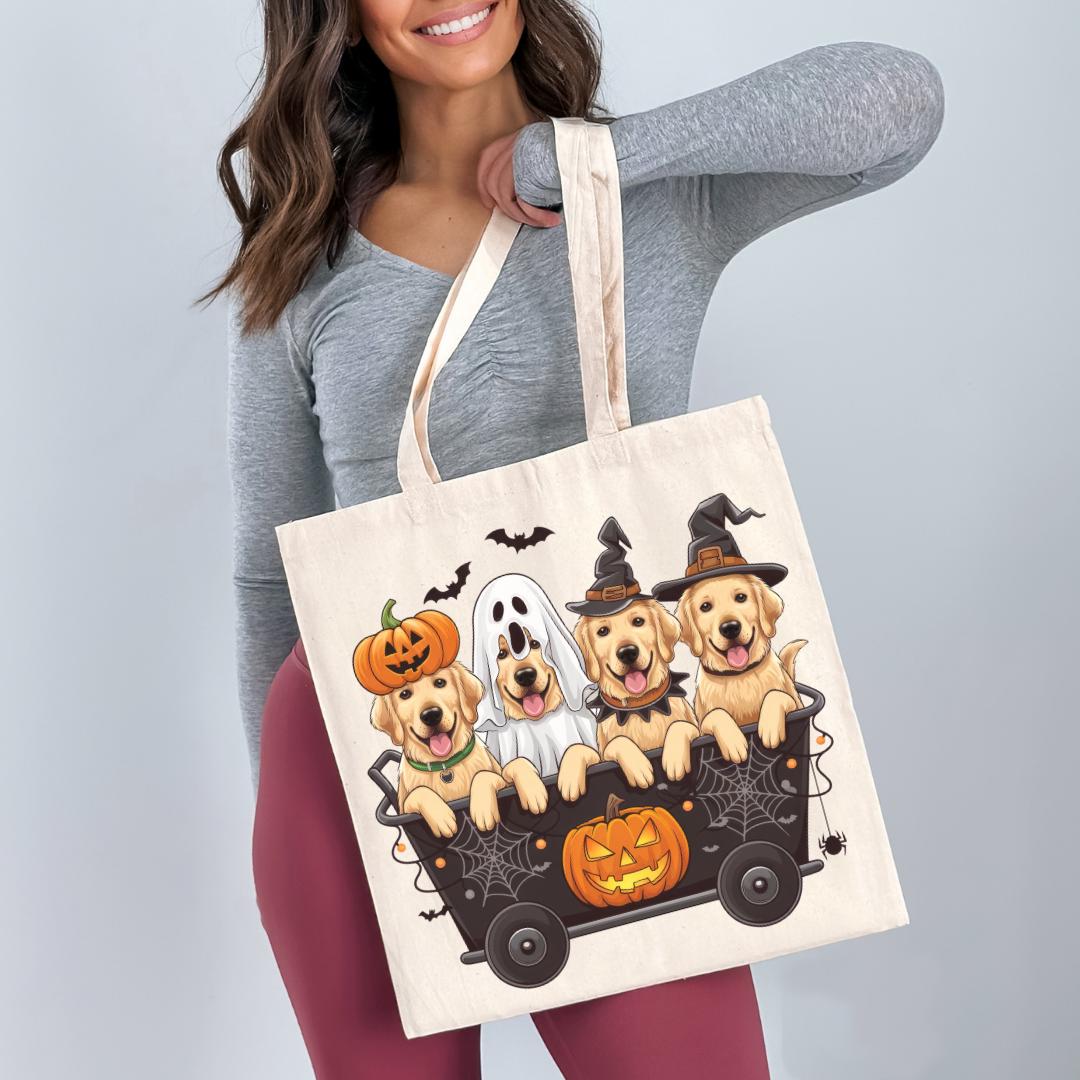 A woman is holding a tote bag with a Halloween-themed illustration of four dogs in a wagon, dressed in costumes and sitting on a pumpkin carriage.