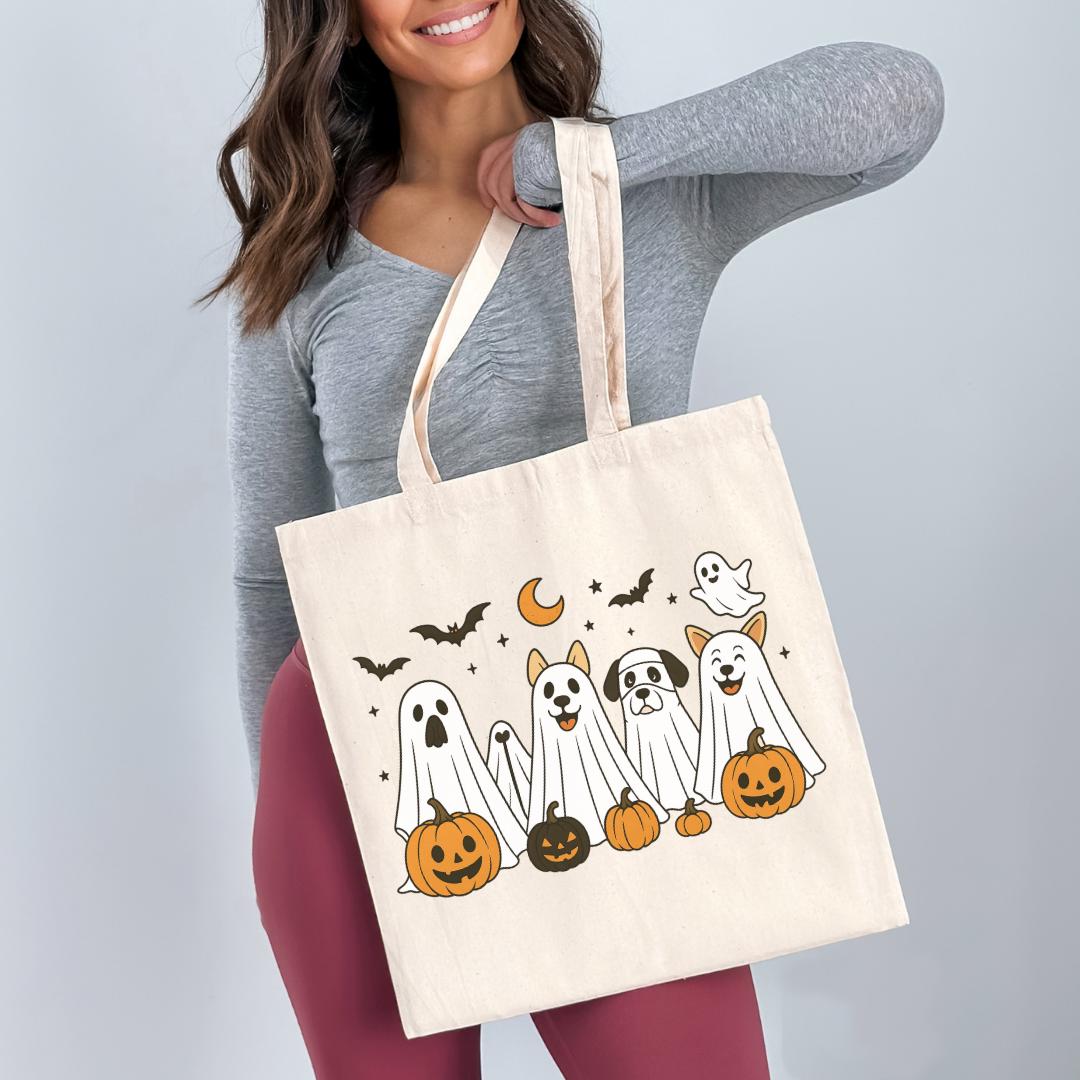 A woman is holding a white tote bag with a Halloween-themed design featuring ghostly figures, pumpkins, and bats.