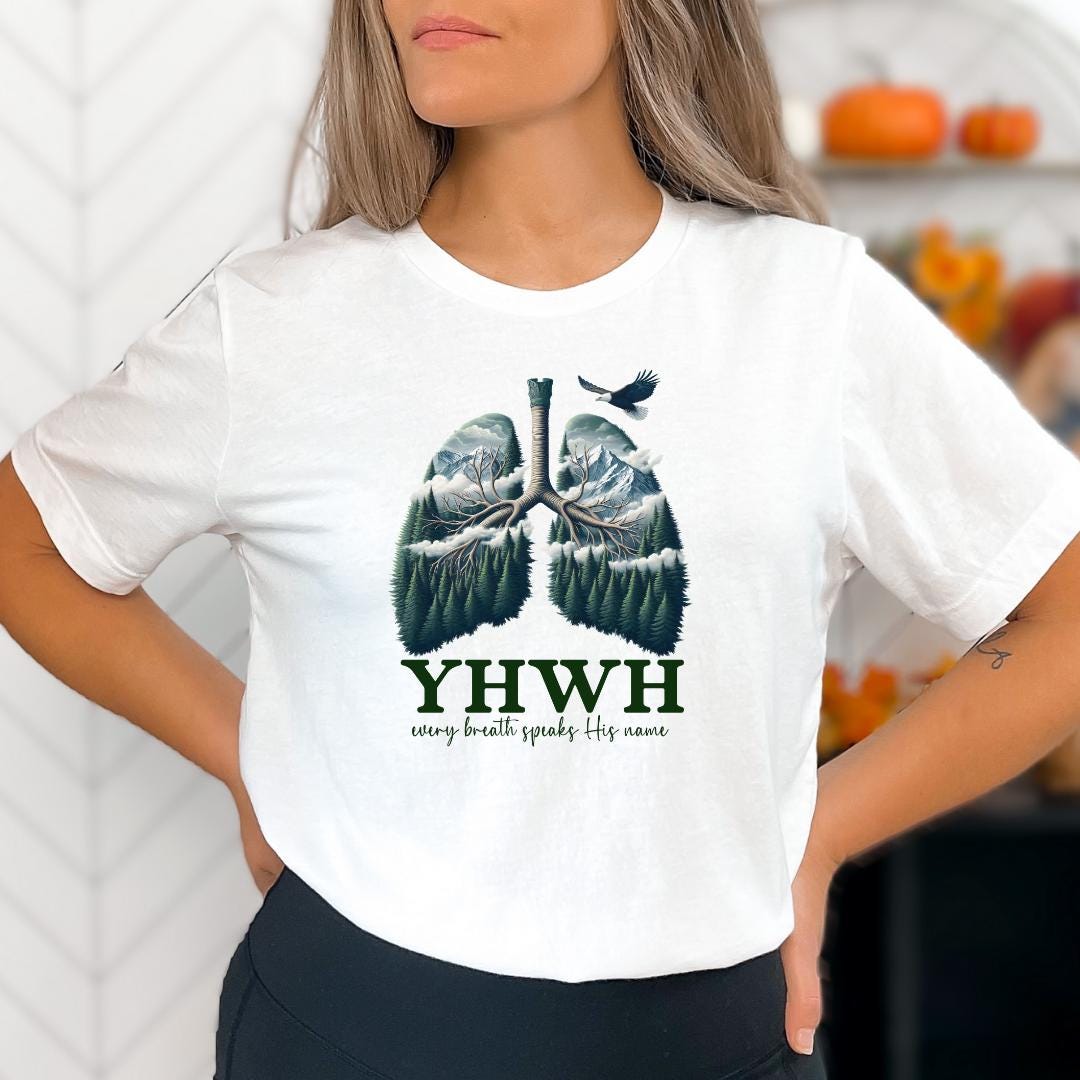 A woman wearing a white t-shirt with a graphic design of lungs and mountains, along with the text "YHWH every breath speaks His name".