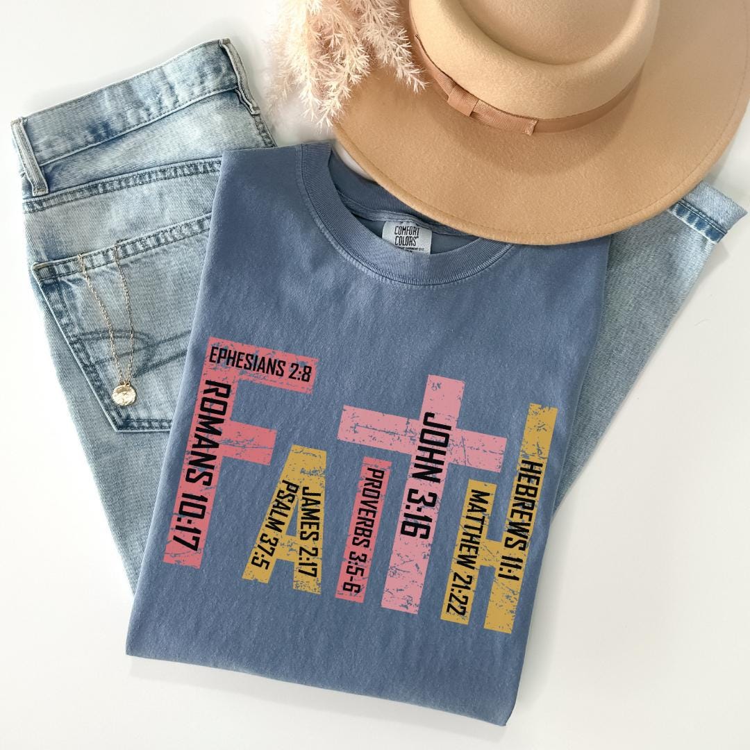 A blue t-shirt with text printed on it, along with a pair of jeans and a beige hat, all laid out on a white surface.