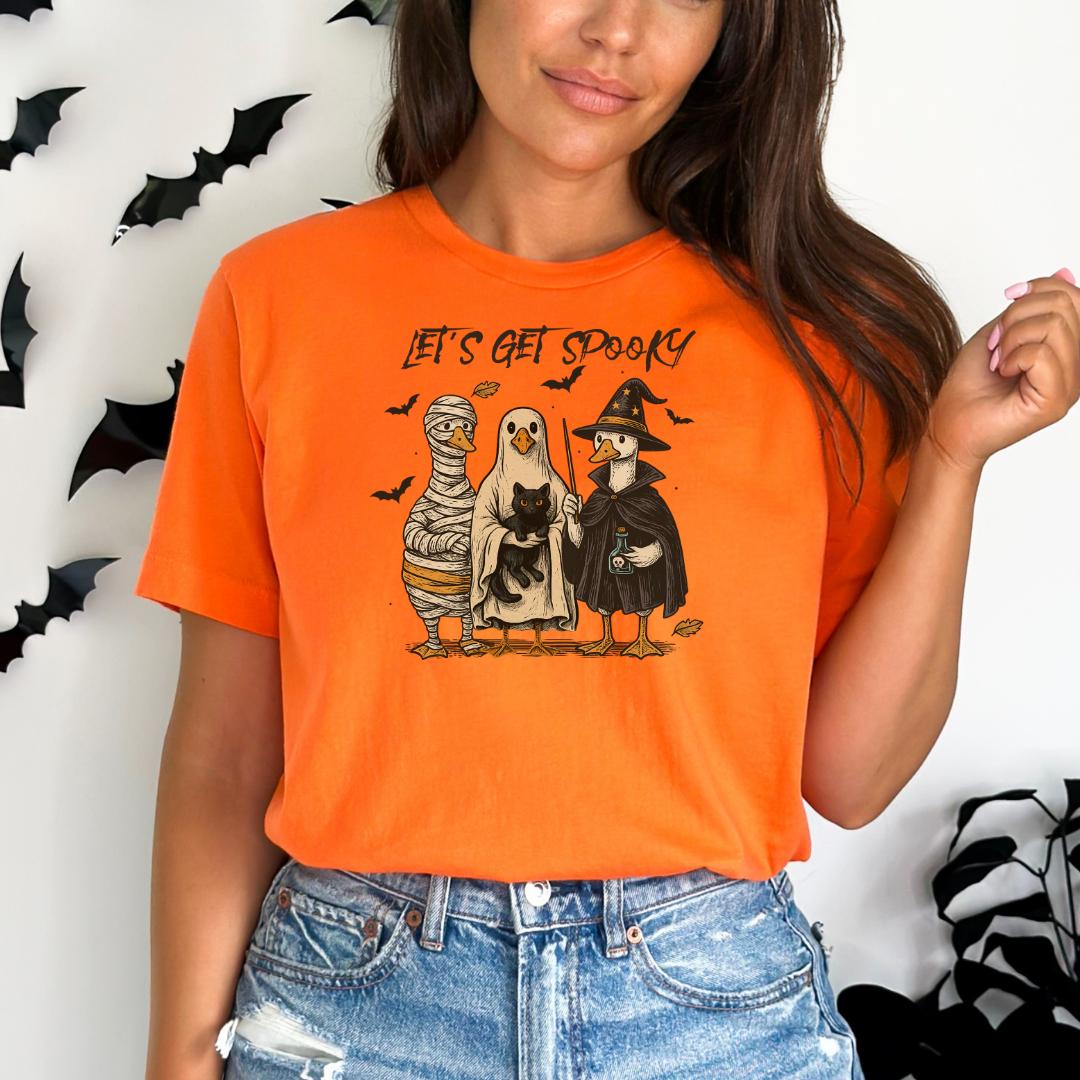 A woman wearing an orange t-shirt with a Halloween-themed graphic of three cartoon characters: a ghost, a mummy, and a witch.