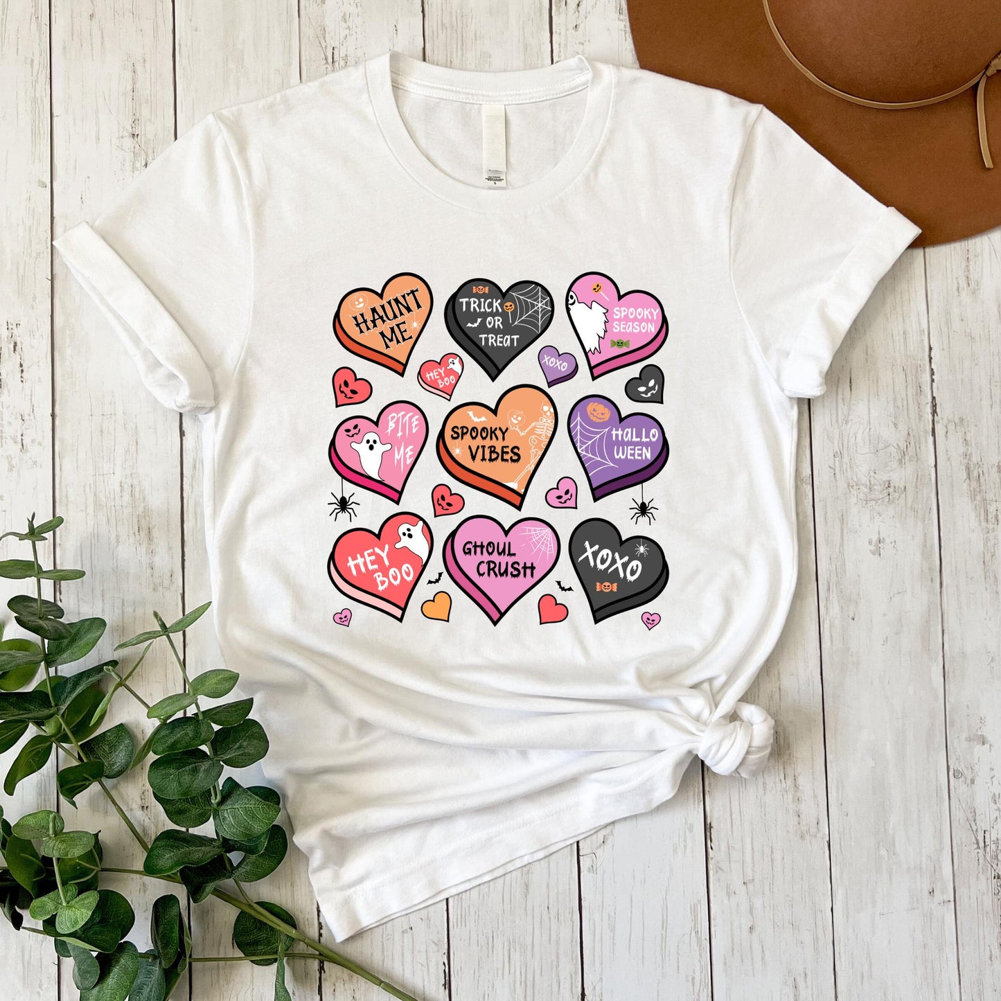 A white t-shirt with a graphic design featuring various Halloween-themed hearts and text, including phrases like "Haunt me", "Trick or treat", "Spooky vibes", "Hollie crush", and "XOXO". The t-shirt is placed on a wooden surface, with a brown hat visible in the background.